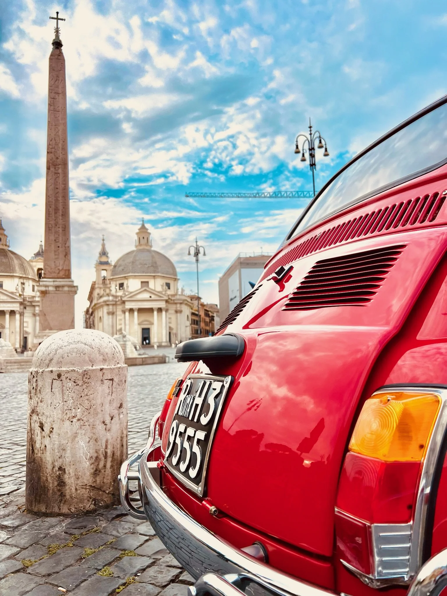Auto rossa parcheggiata su una strada di ciottoli in una piazza storica con edifici antichi e un obelisco, cielo azzurro con nuvole