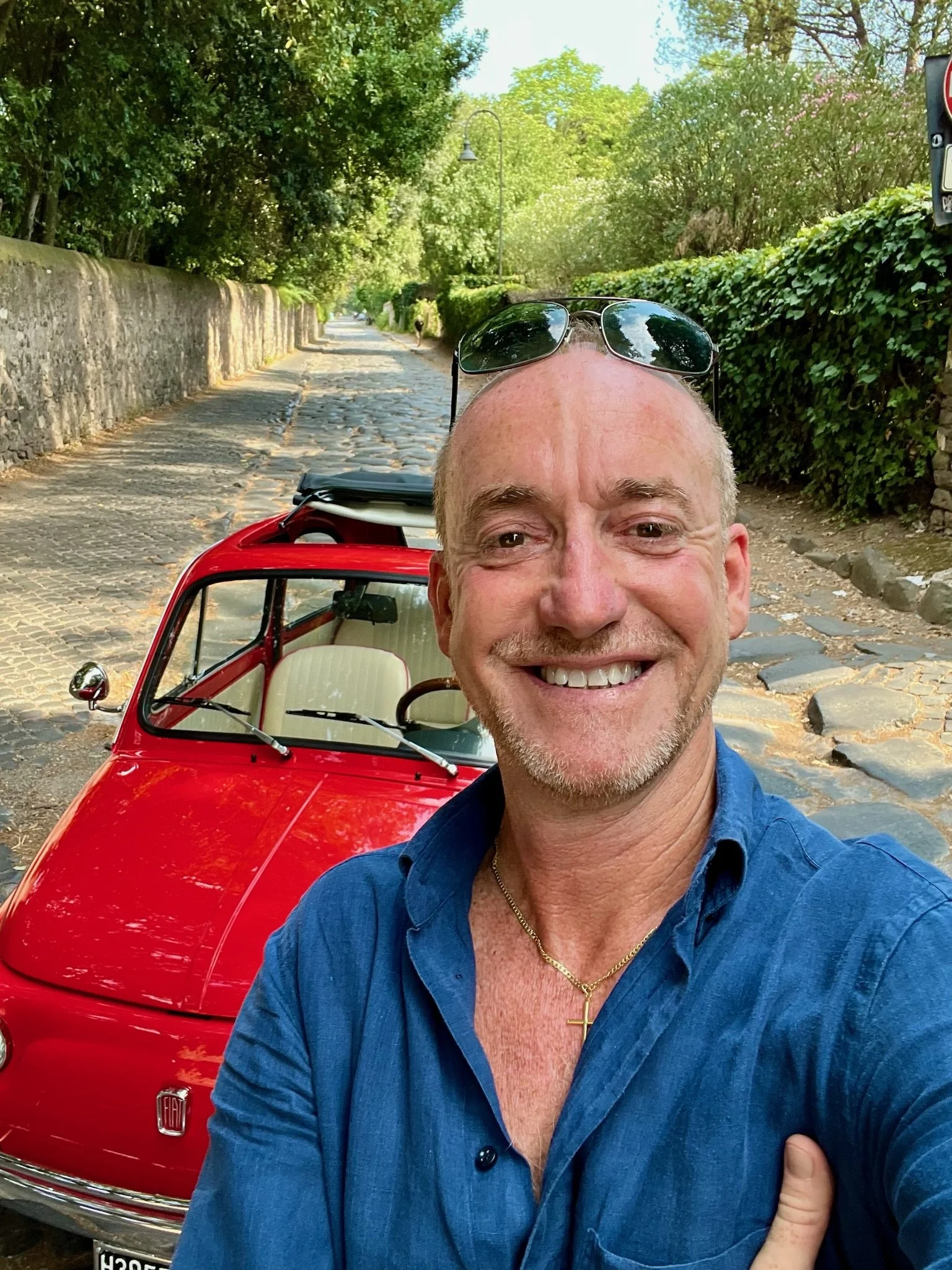 Uomo sorridente con occhiali da sole sulla testa, che indossa una camicia blu, che si prende un selfie davanti a una macchina rossa cabrio, su una strada pavimentata con ciottoli, circondato da alberi e cespugli verdi.