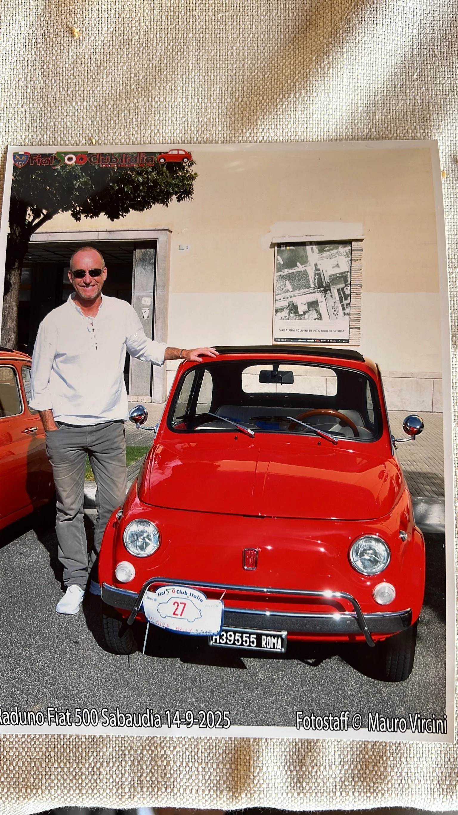 Un uomo sorridente con occhiali da sole indossa una camicia bianca e pantaloni grigi, poggia una mano su un'auto rossa Fiat 500 davanti a un edificio, durante un raduno di auto d'epoca il 14 settembre 2025 a Sabaudia.