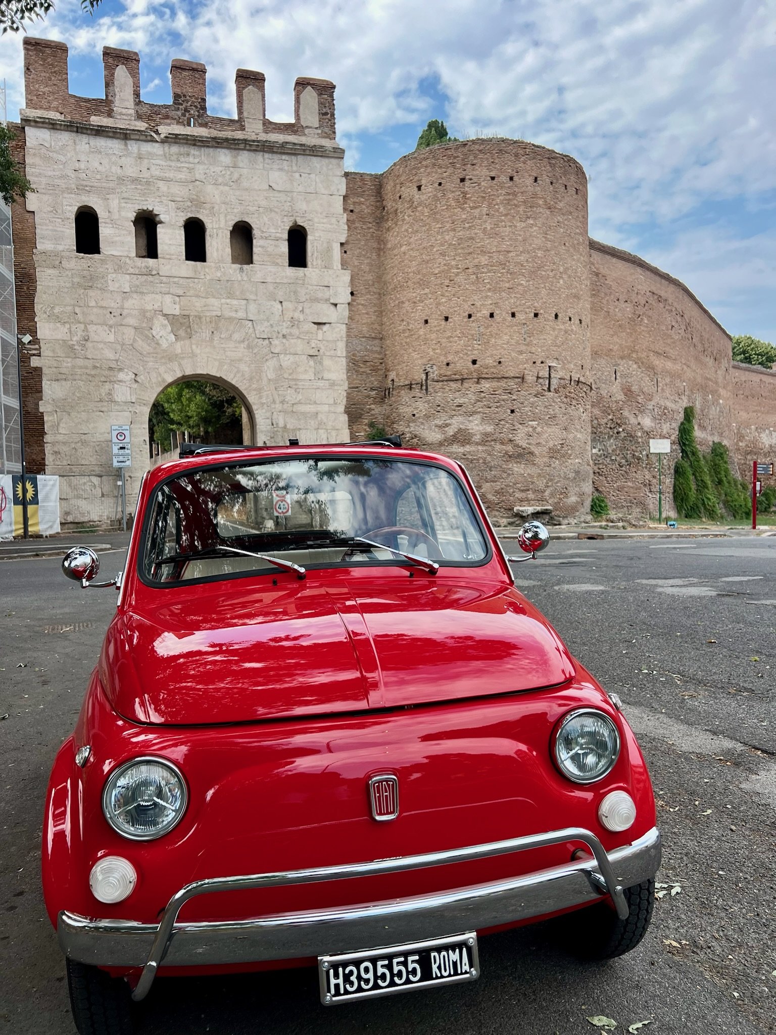Auto d'epoca rossa parcheggiata davanti a una porta di pietra e un'alta mura in mattone, con un cielo nuvoloso sopra Roma.