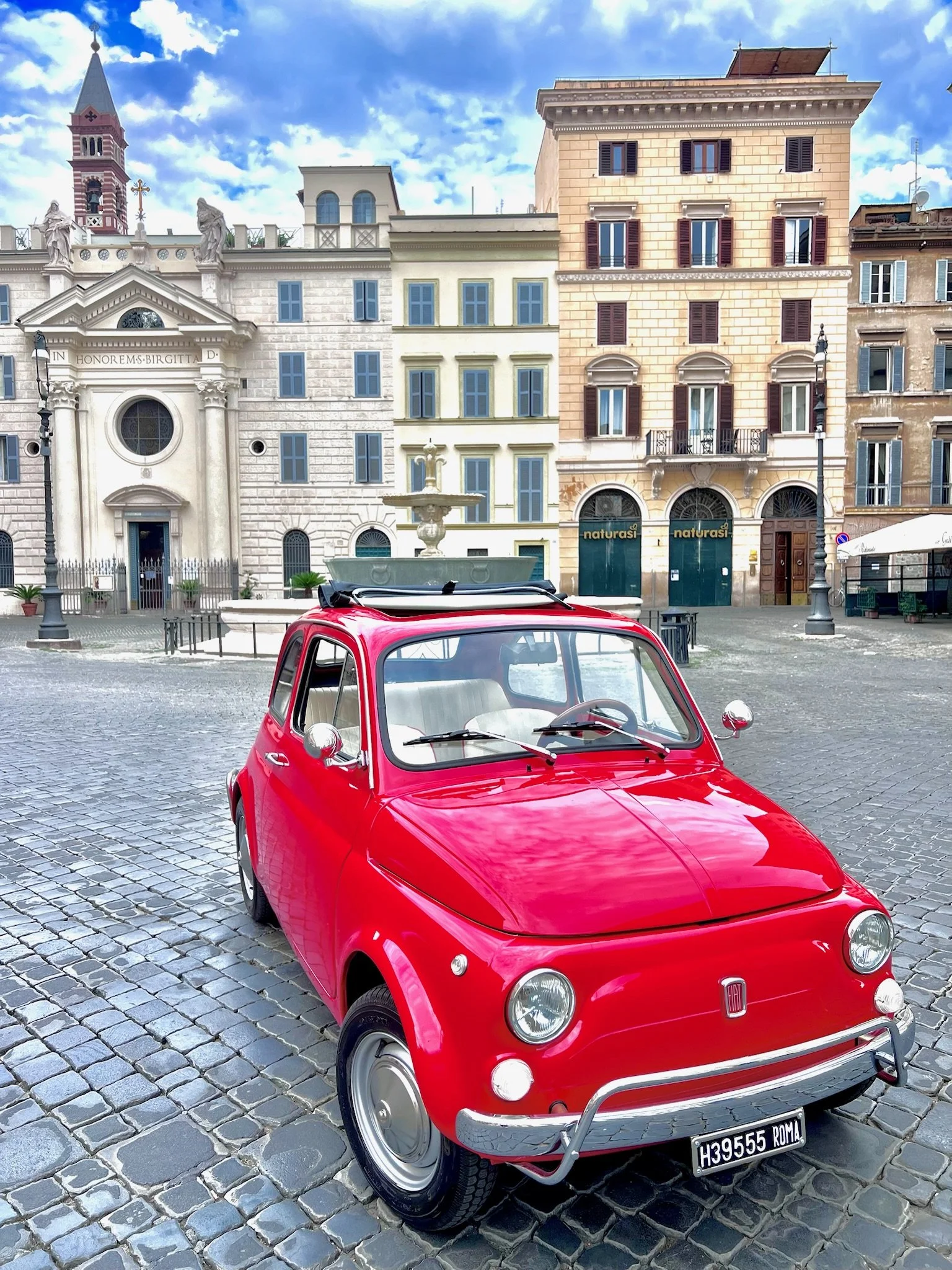 Auto rossa Fiat 500 parcheggiata su una piazza con edifici storici e fontana sullo sfondo, cielo nuvoloso.