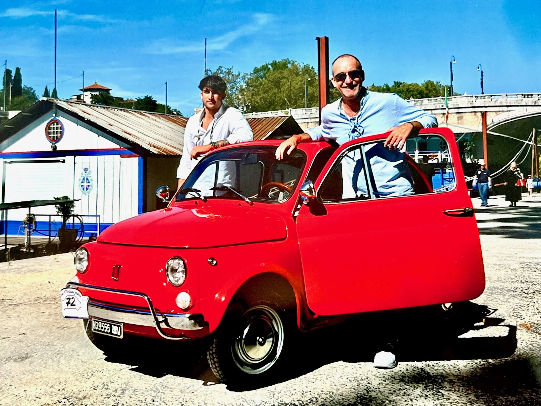 Due uomini con cappelli e occhiali da sole sono vicino a un'auto rossa vintage a forma di quadriciclo, sotto un cielo azzurro. Uno di loro è seduto sul tetto e l'altro vicino davanti.