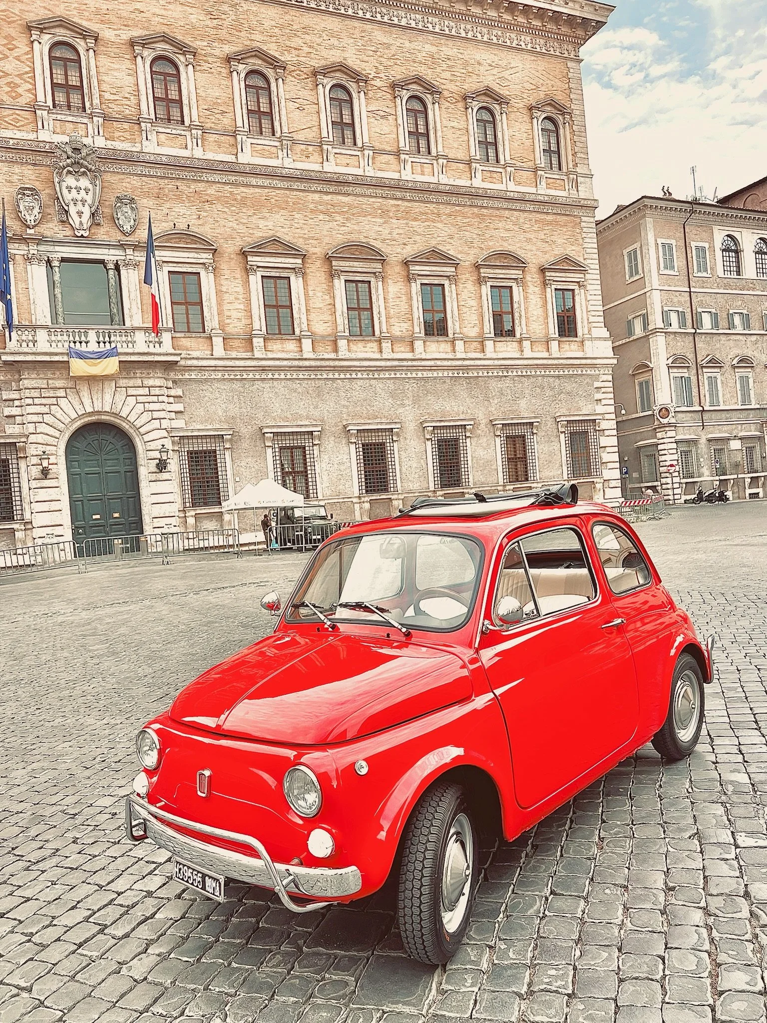 Auto d'epoca rossa parcheggiata su una piazza di pietra davanti a un grande edificio storico con bandiere.