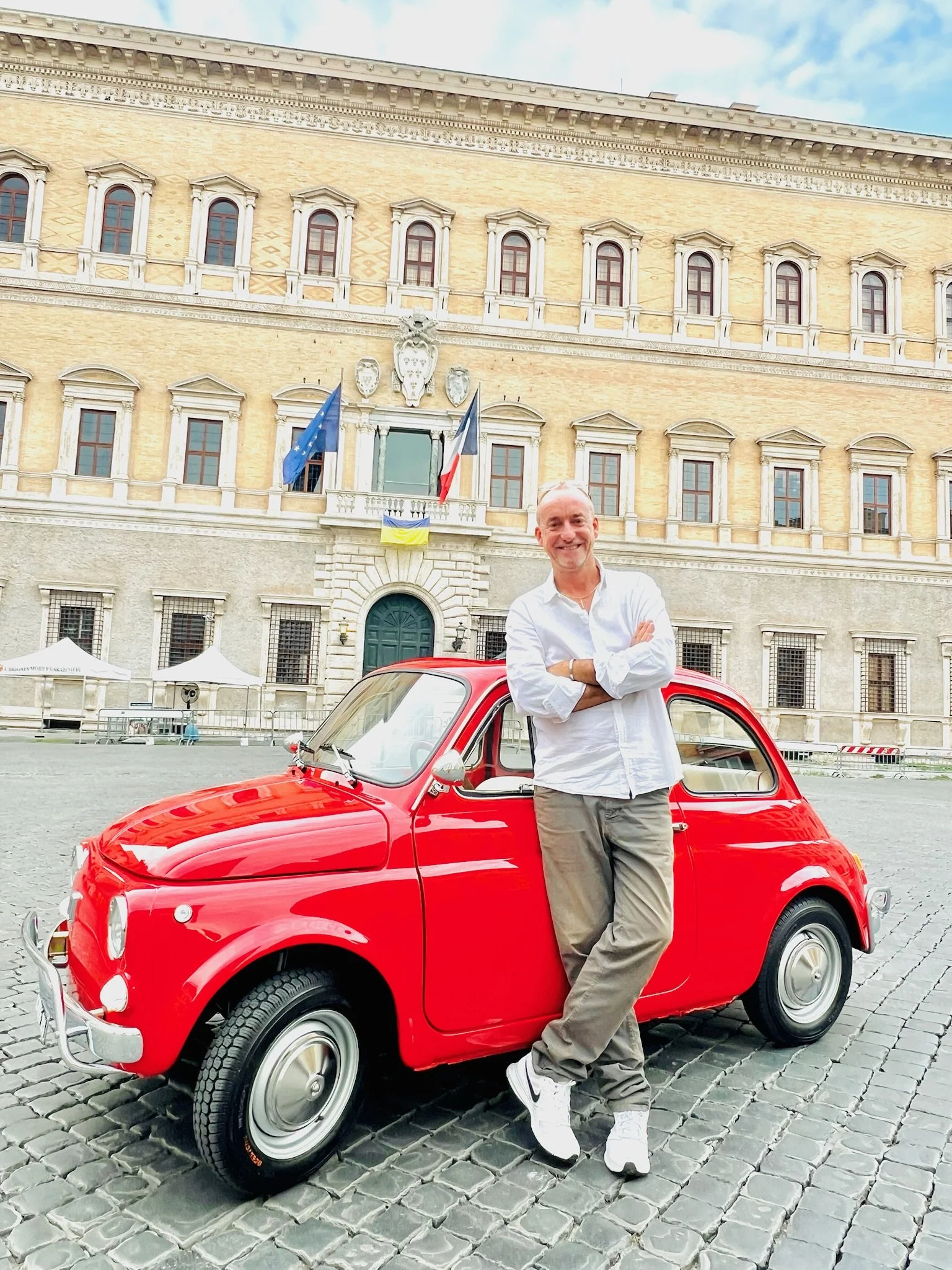 Uomo sorridente che si appoggia a una piccola auto rossa, davanti un edificio storico con bandiere, in piazza con pavimentazione in pietra.