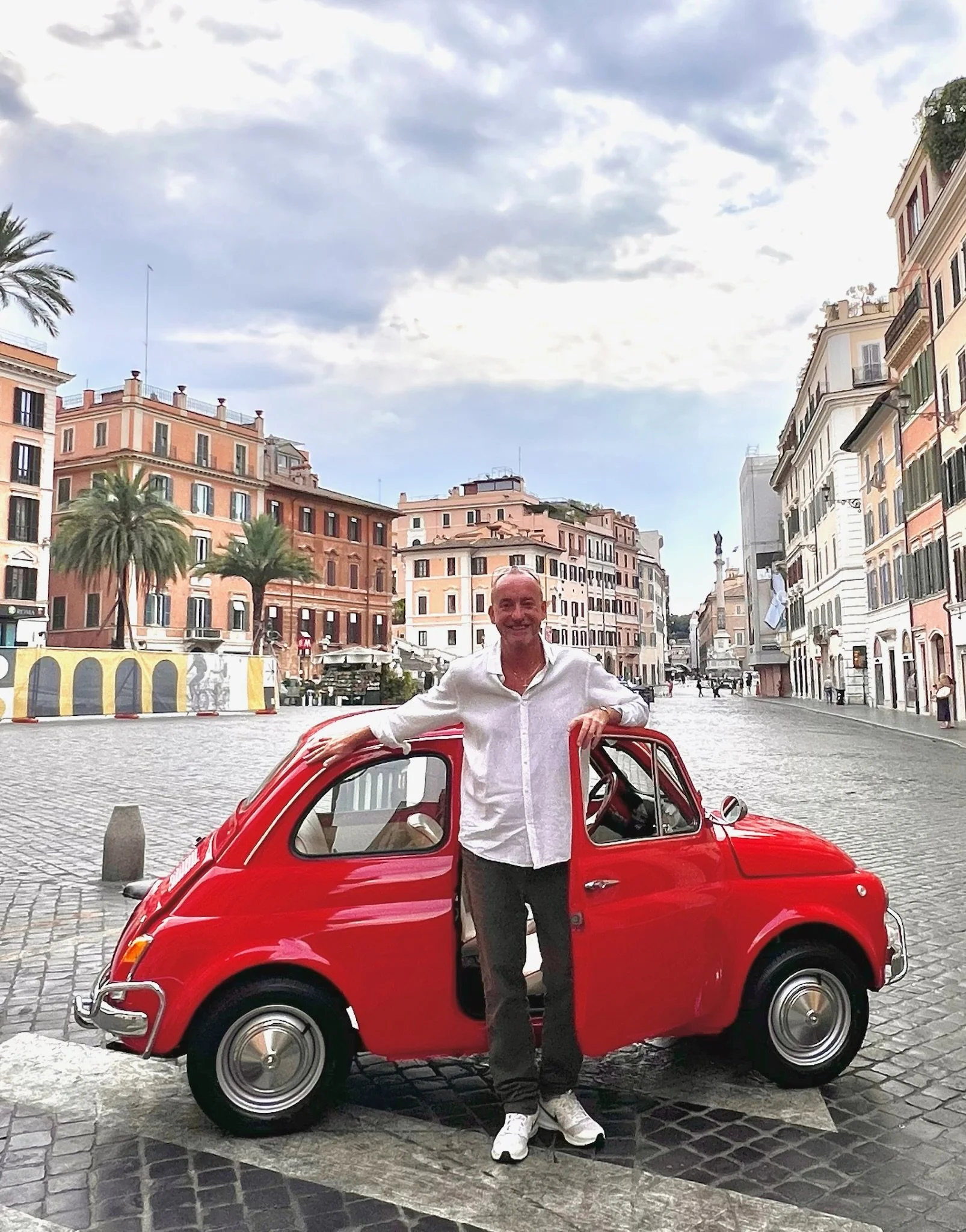Uomo sorridente con camicia bianca e pantaloni scuri si appoggia su una piccola auto rossa in una piazza di città con edifici colorati e palme sullo sfondo.