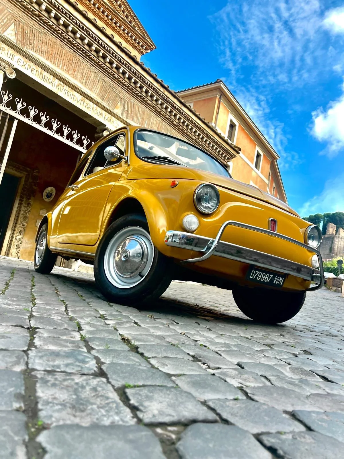 Auto d'epoca di colore giallo in primo piano con sfondo di edificio storico in pietra e cielo blu con nuvole