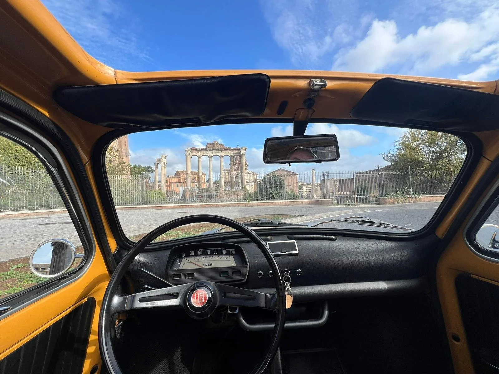 Interno di una vecchia auto gialla con quadro strumenti e volante, vista attraverso il parabrezza con rovine romane antiche e cielo azzurro con nuvole