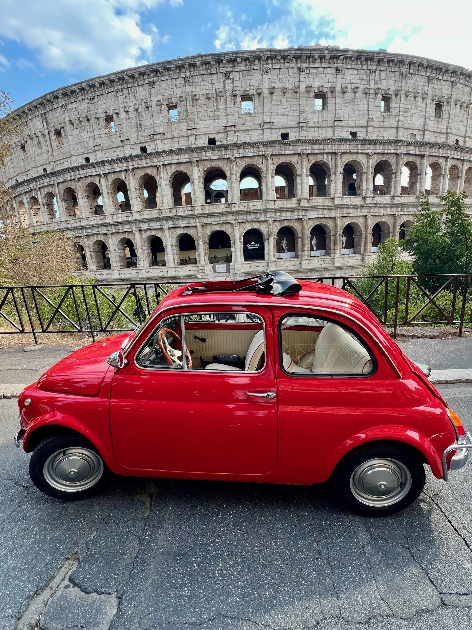 Piccolo auto rosso parcheggiato davanti al Colosseo a Roma, con un cielo parzialmente nuvoloso sopra.