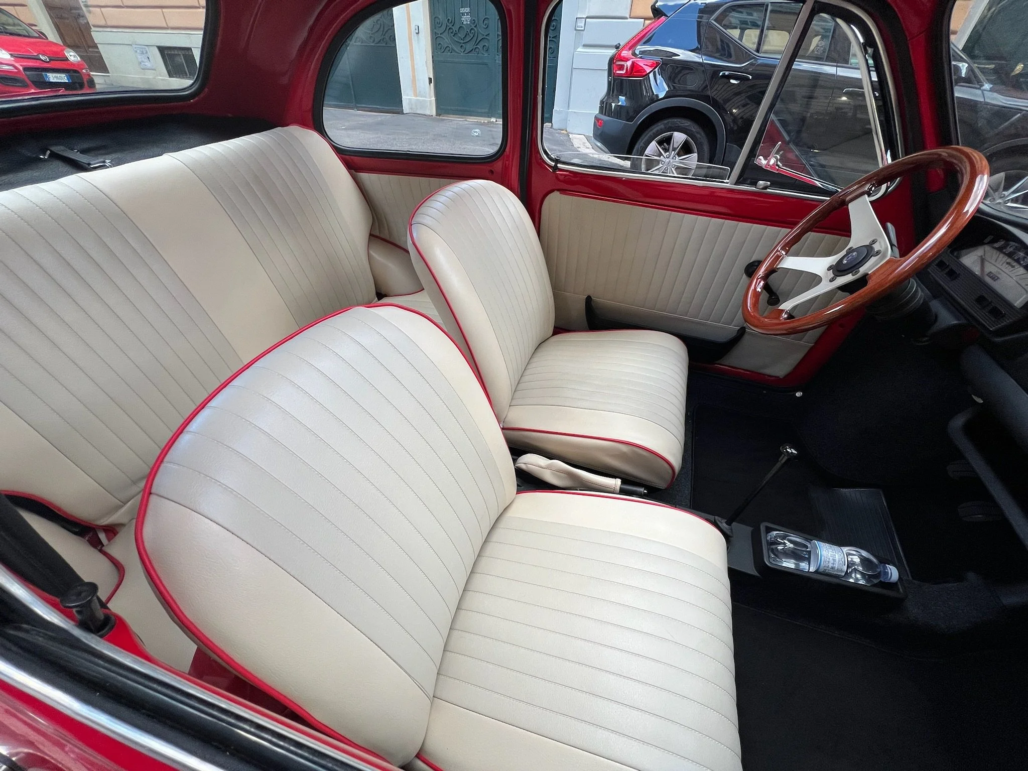 Interno di una vettura vintage con sedili beige con finiture rosse, volante in legno e cruscotto nero.