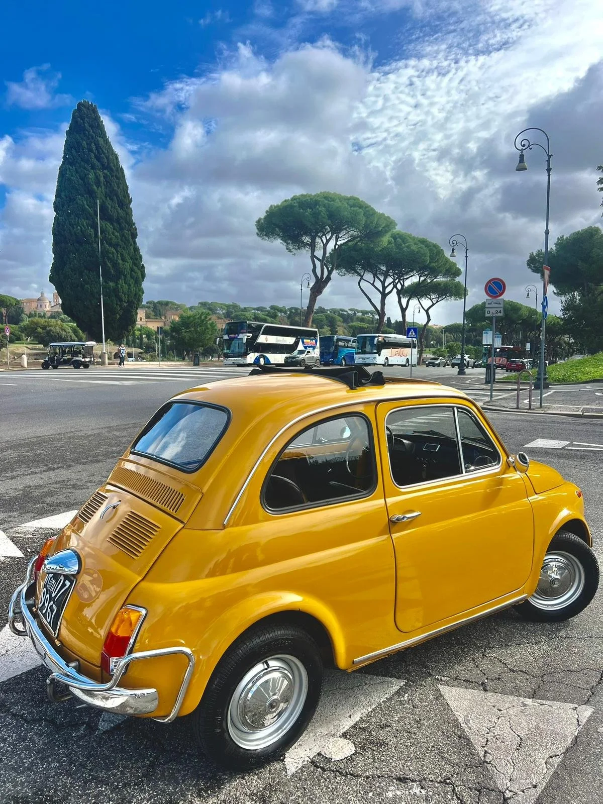 Auto d'epoca gialla parcheggiata in una strada con alberi, autobus e lampioni sullo sfondo, cielo nuvoloso.