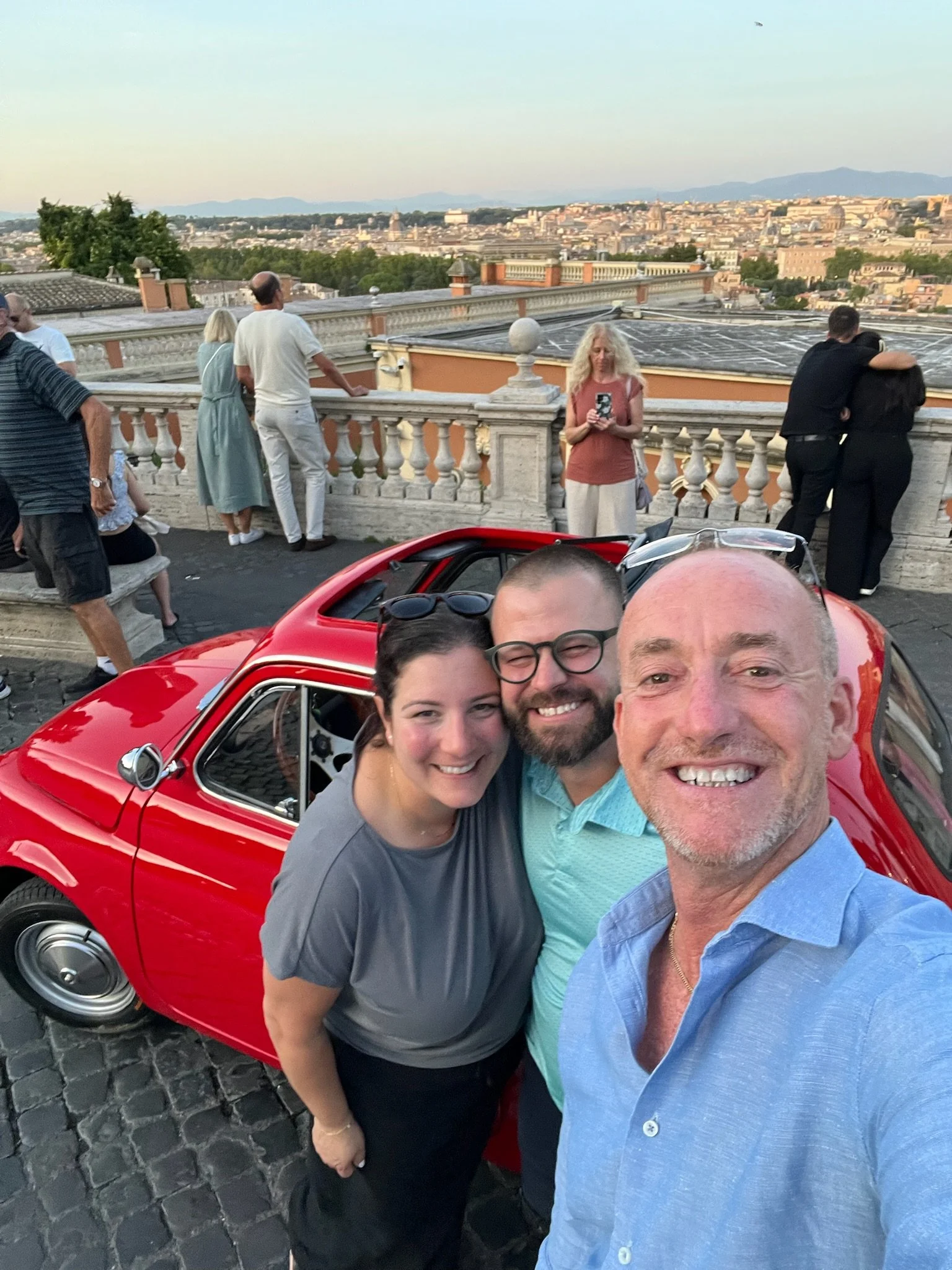 Un gruppo di persone si stavano facendo una foto davanti a una vista panoramica di una città, con una macchina rossa in primo piano. Sullo sfondo, persone che guardano la città e scattano foto, in una zona con un parapetto ornamentale.