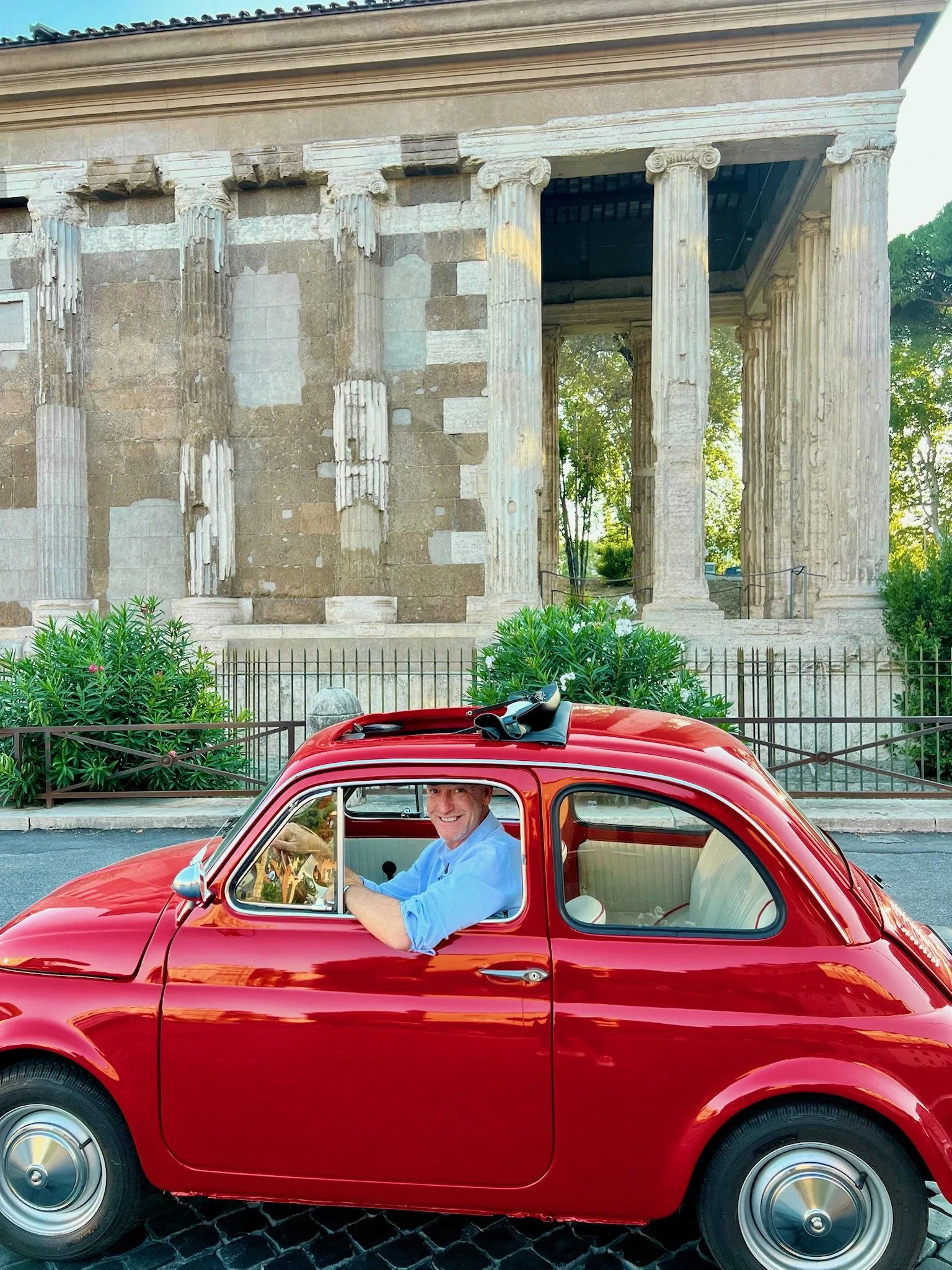 Uomo seduto in un'auto rossa vintage con monumento greco antico sullo sfondo.