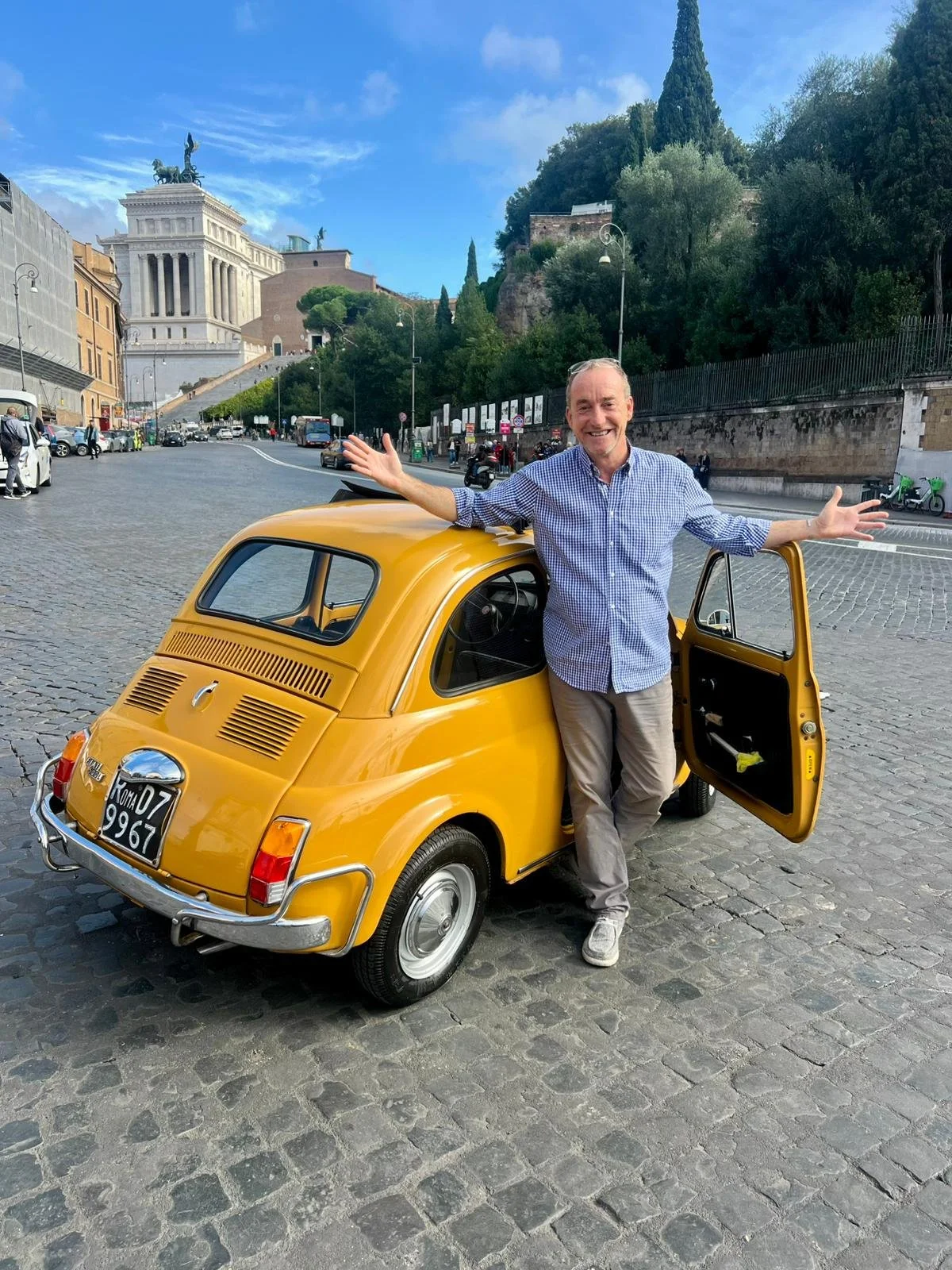 Uomo sorridente con braccia aperte accanto a una piccola auto gialla in una piazza di Roma, con scalinata e statue sullo sfondo.