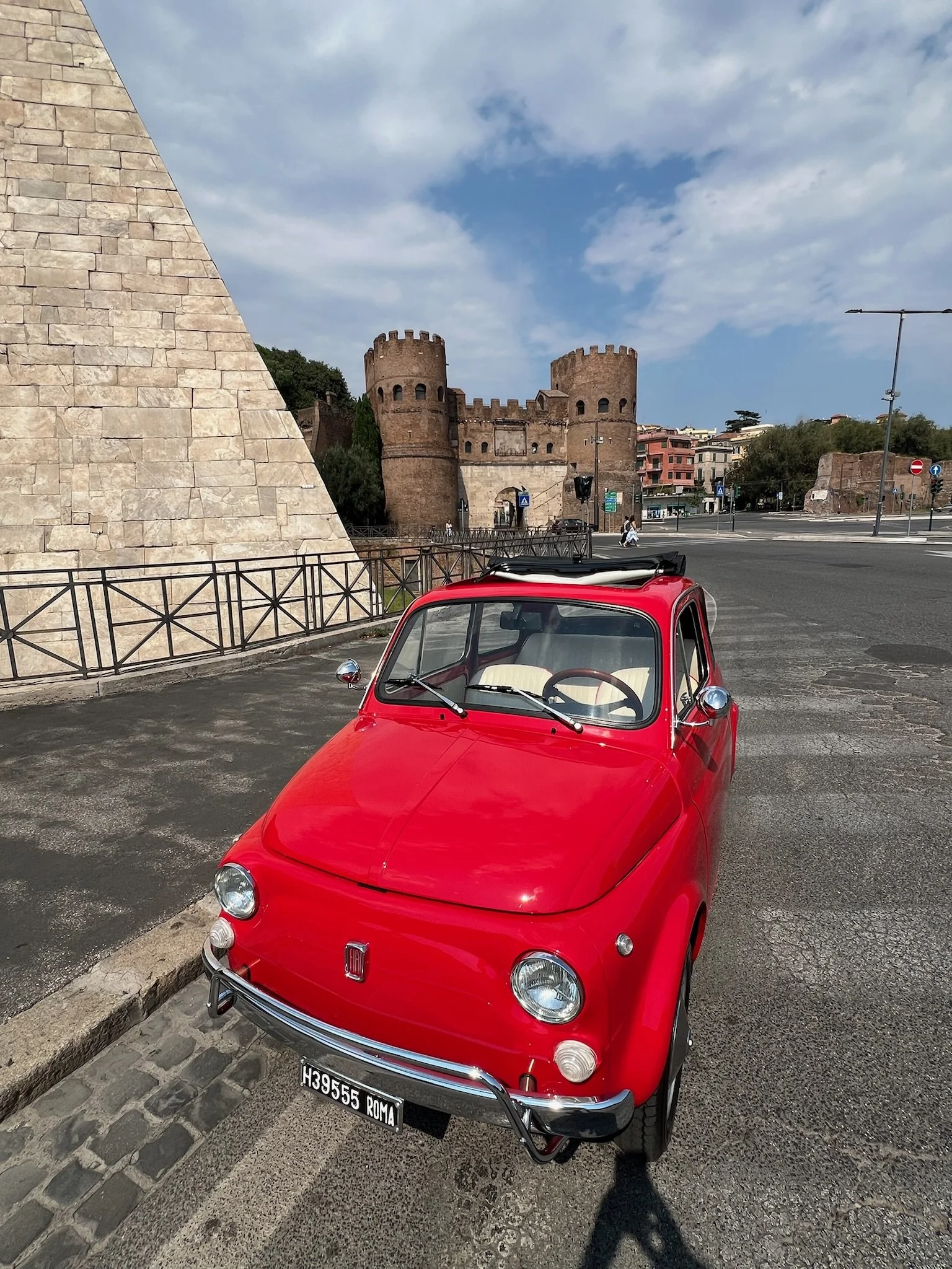 Auto rossa Fiat 500 parcheggiata in strada di fronte a un castello in pietra con torri circolari, sullo sfondo edifici e un cielo parzialmente nuvoloso.