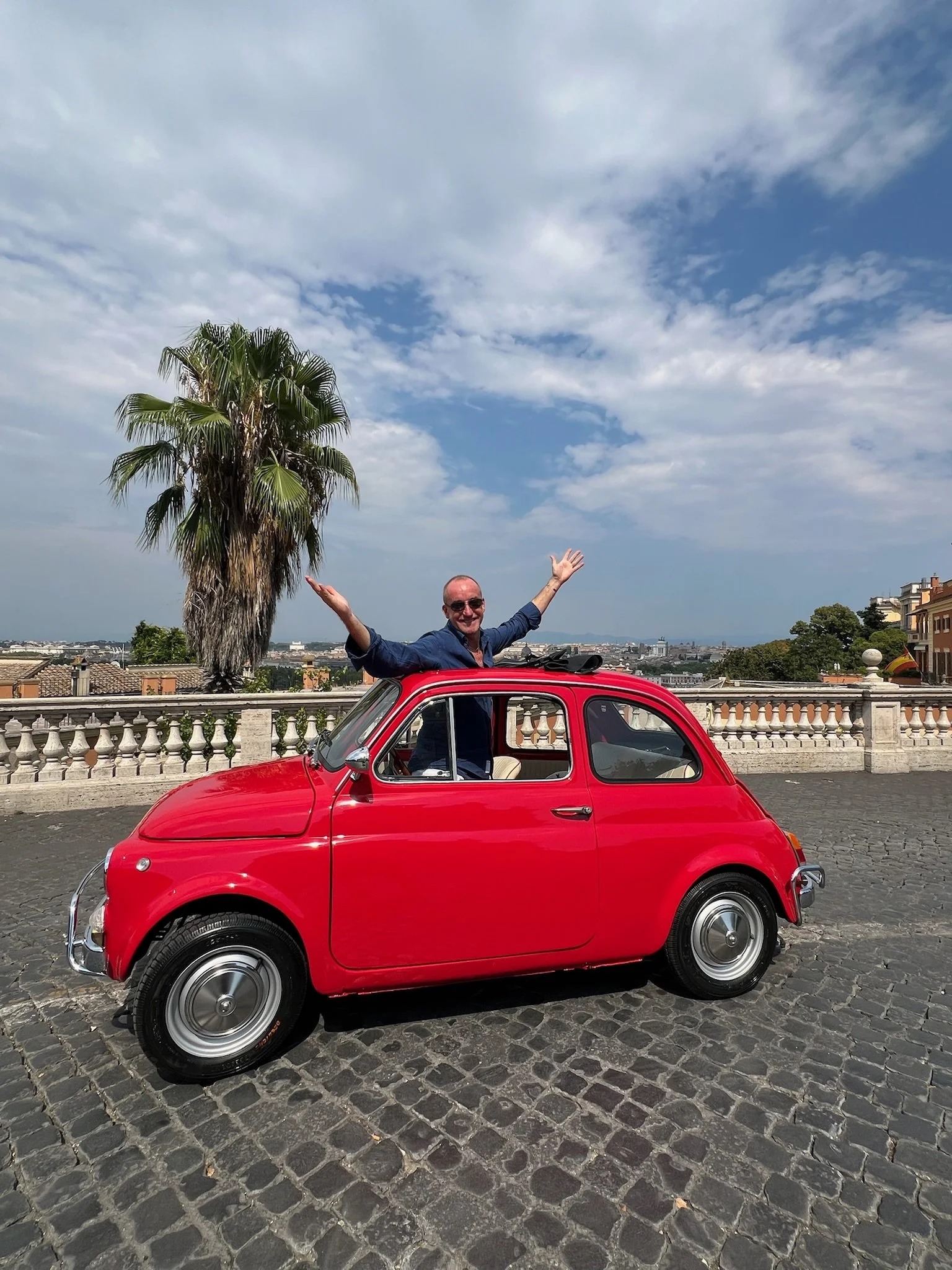 Uomo con occhiali che sorride con le braccia aperte sopra una piccola auto rossa parcheggiata su una strada di ciottoli, con un albero di palma e un cielo nuvoloso sullo sfondo.