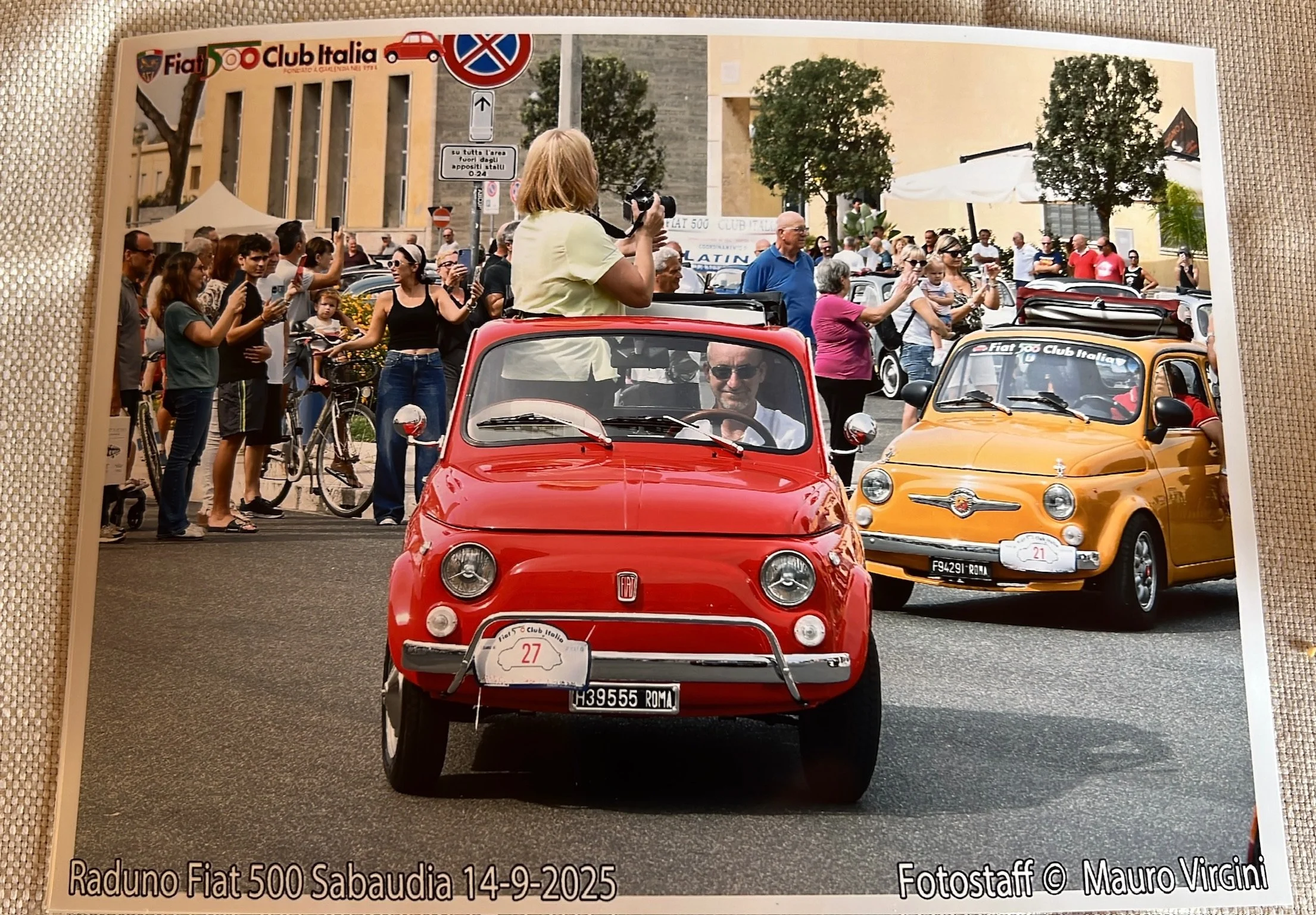 Un raduno di automobili Fiat 500 a Sabaudia con molte persone che osservano e fotografano le vetture, un'auto rossa in primo piano con un conducente e una donna in piedi che le scatta una foto.