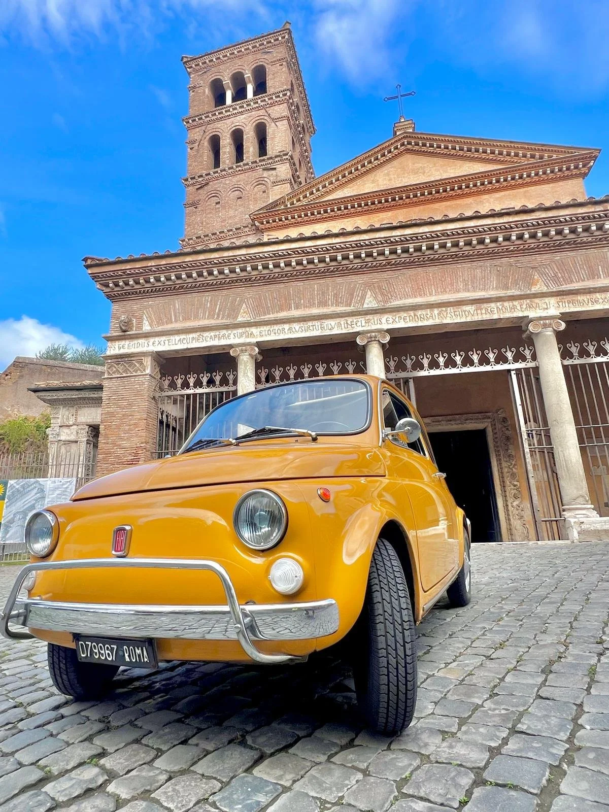 Auto Fiat giallo d'epoca parcheggiato davanti a una chiesa in pietra a Roma, con un cielo blu e alcune nuvole.