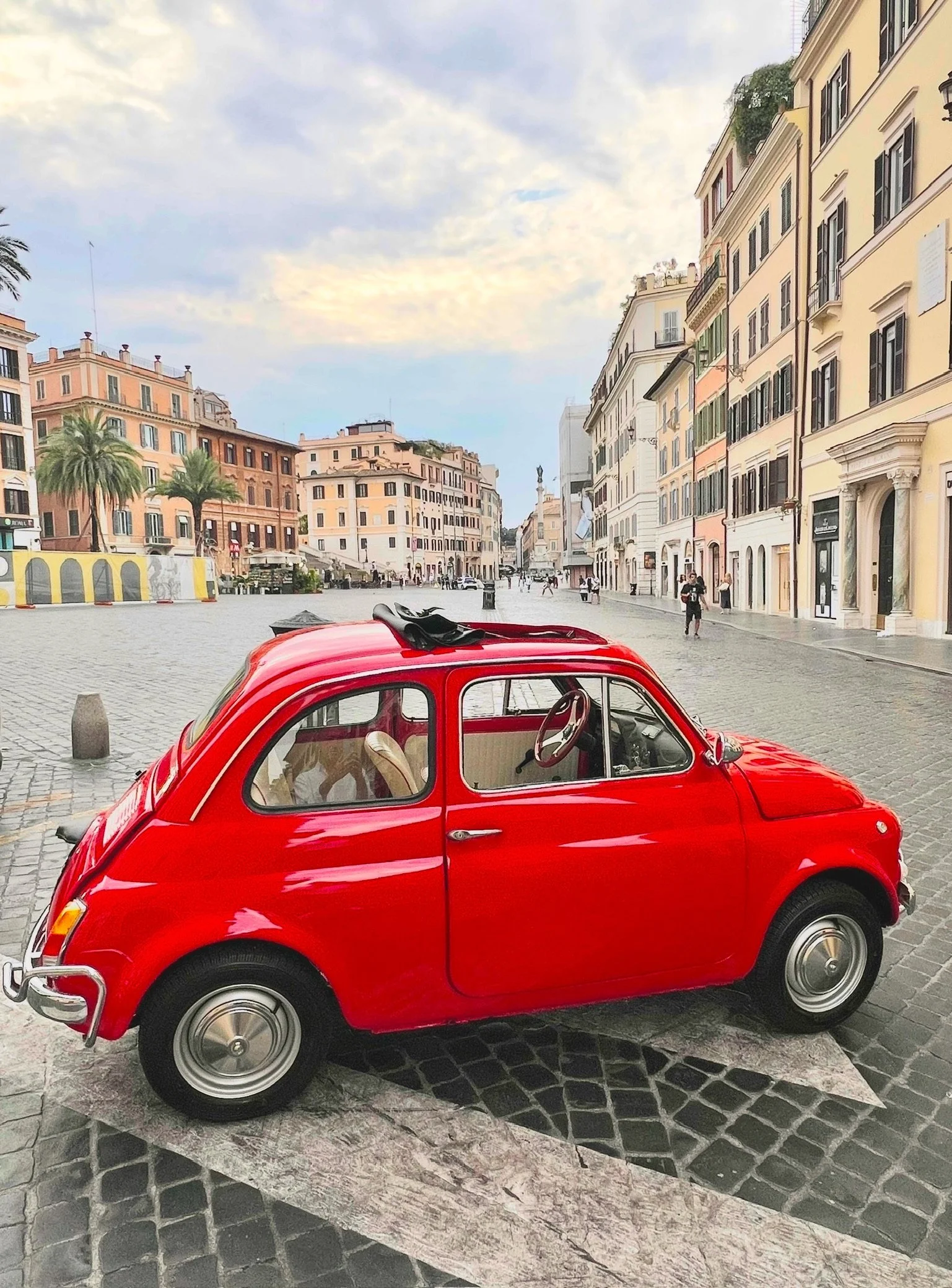 Una mini macchina rossa parcheggiata su una piazza con edifici colorati e alcuni palmizi sullo sfondo.
