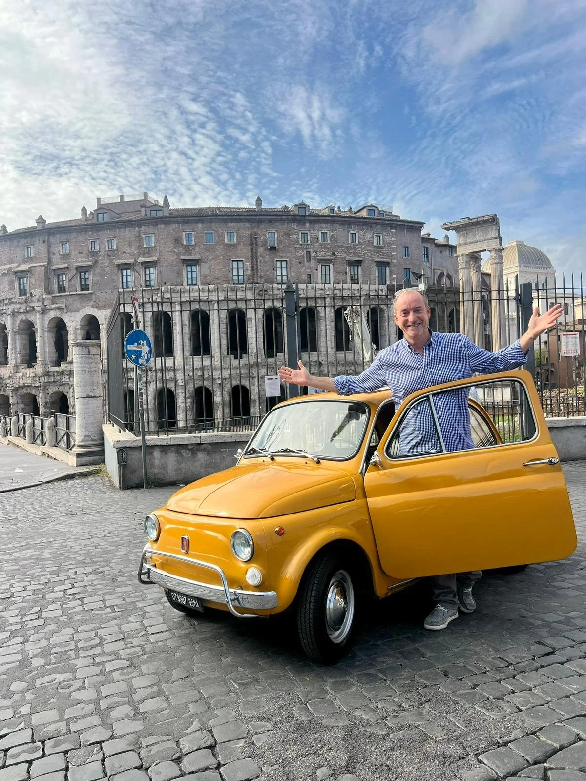 Uomo felice con braccia aperte che si trova accanto a una piccola auto gialla, con all'interno tutto il corpo visibile, in una strada di ciottoli davanti alle rovine dell'Anfiteatro Flavio a Roma con un cielo blu e nuvole leggere sopra.
