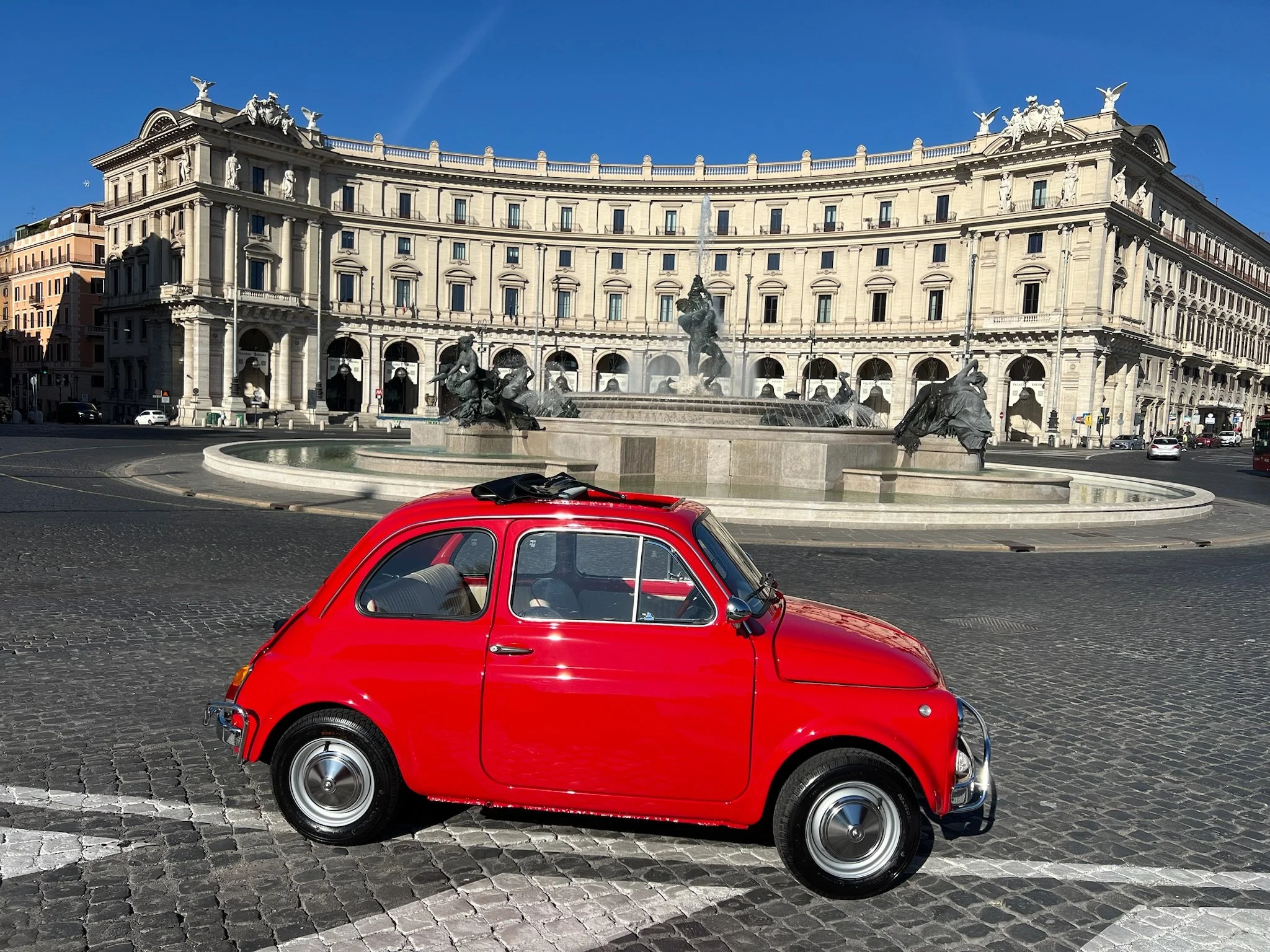 Una piccola auto rossa parcheggiata su una piazza di ciottoli in fronte a una grande fontana con statue di cavalli e figure umane, con un edificio storico sullo sfondo, sotto un cielo blu.