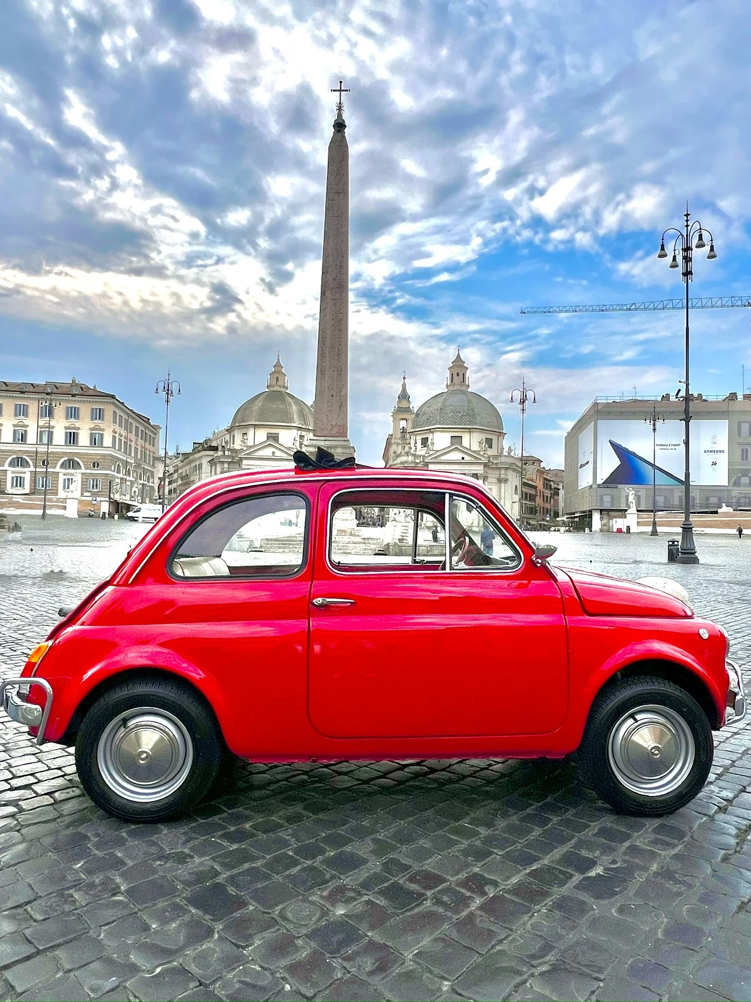 Auto rossa in piazza con obelisco e chiese barocche sullo sfondo, cielo con nuvole