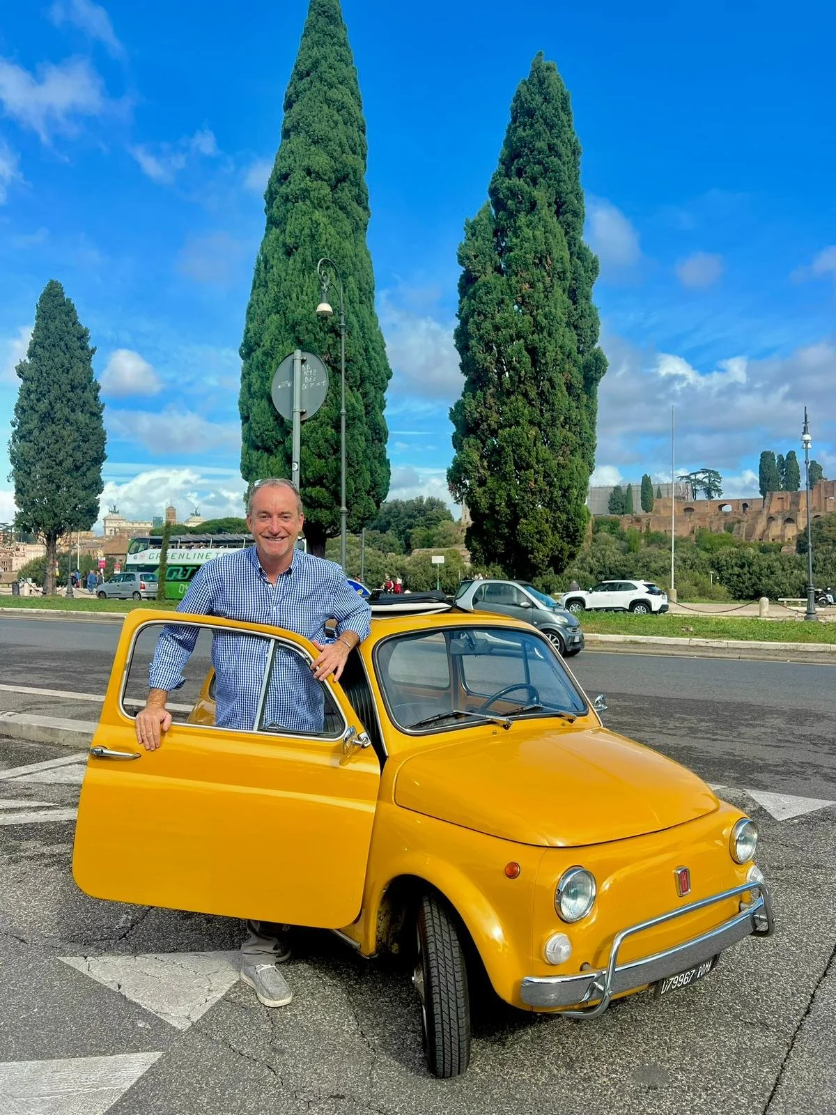 Uomo sorridente che esce da un'auto gialla in un'area urbana con alberi e veicoli in background.