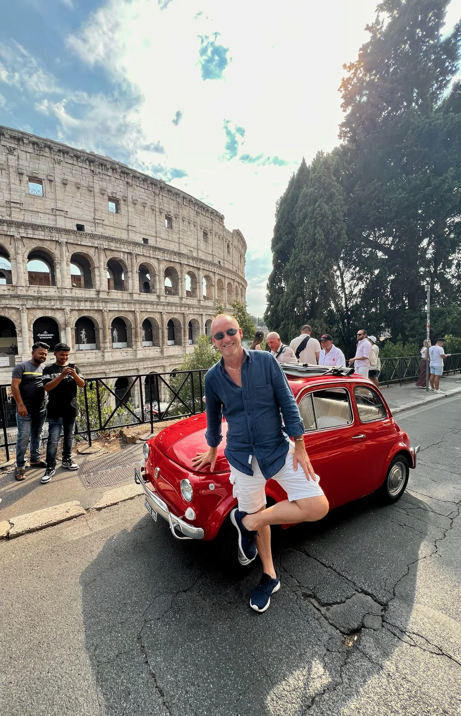 Uomo sorridente con occhiali da sole, vestito con maglietta blu e pantaloncini bianchi, posa accanto a una macchina rossa classica vicino al Colosseo a Roma, con altri turisti sullo sfondo.