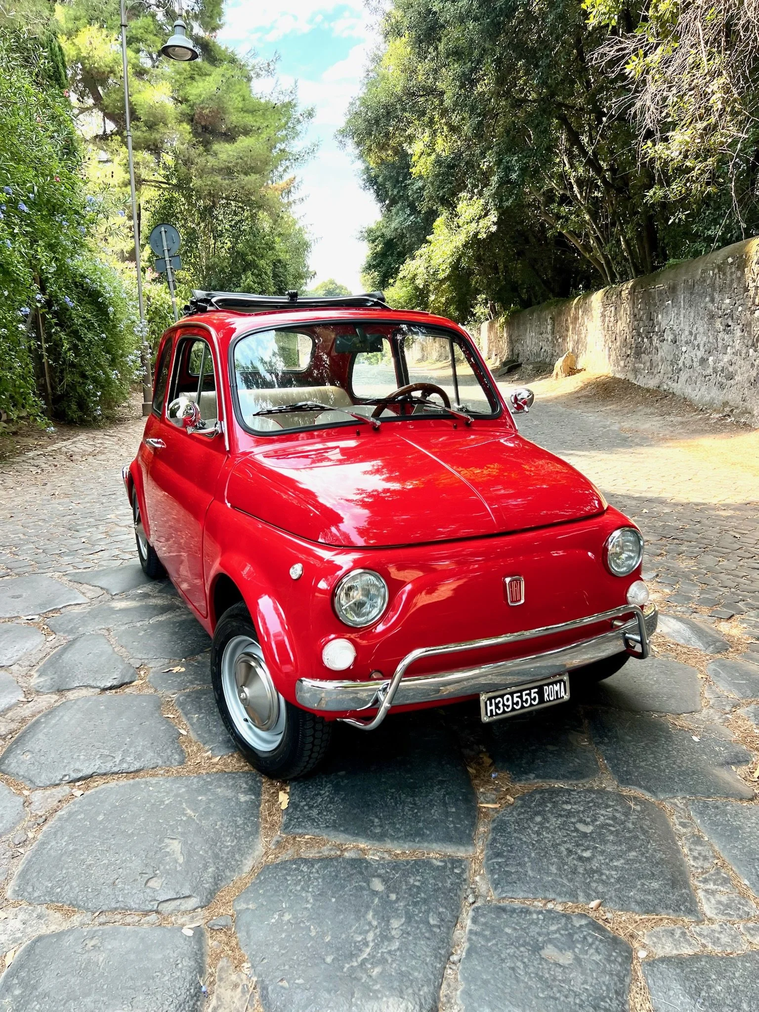 Auto compatto e rosso con copertura sul tetto parcheggiato su una strada laterale di ciottoli, con alberi e muro di pietra sullo sfondo.