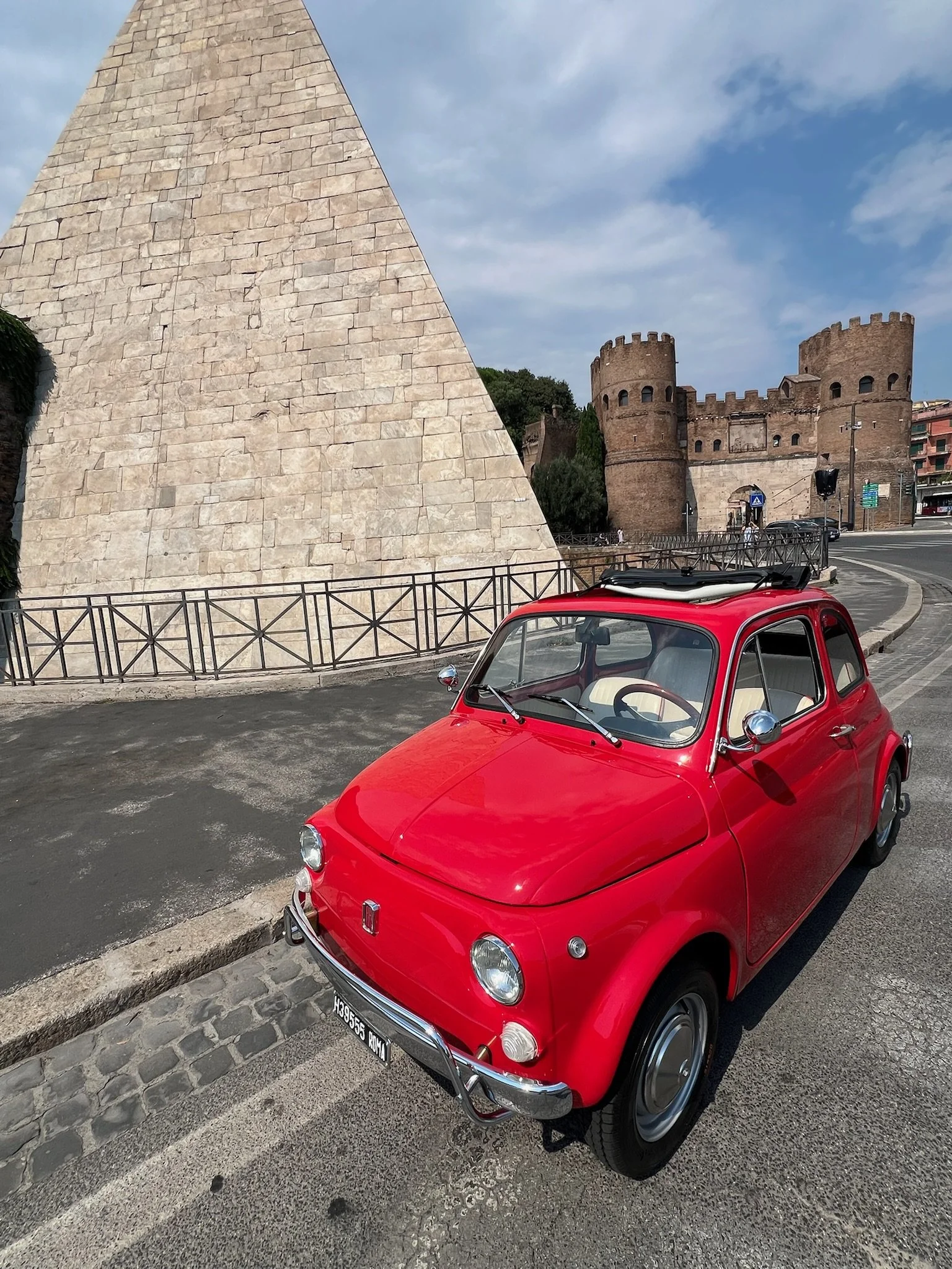 Auto rossa vintage parcheggiata su strada con un castello in pietra e una struttura piramidale in pietra alle spalle, sotto un cielo parzialmente nuvoloso.