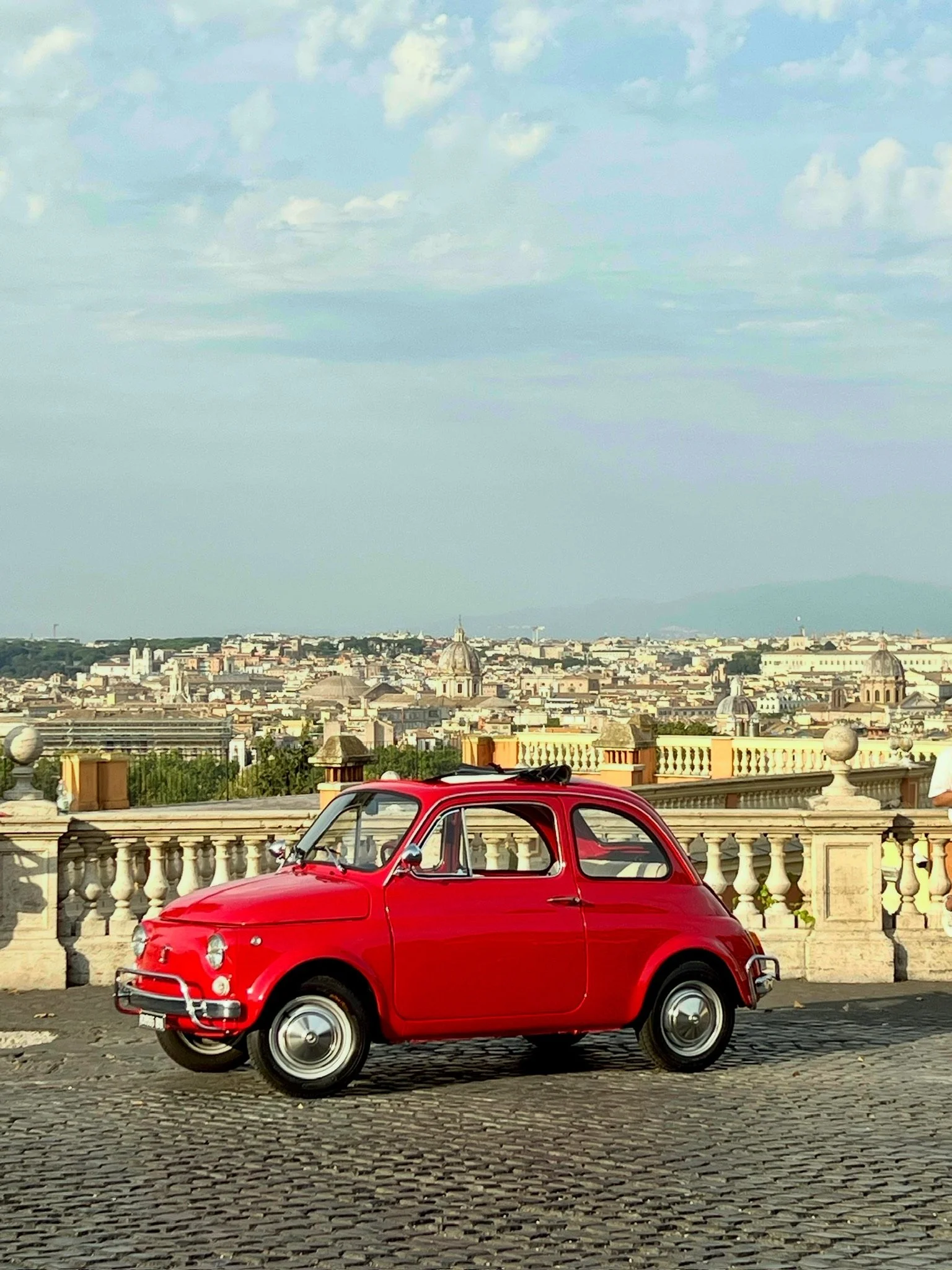 Una piccola macchina rossa è parcheggiata su un muretto con una vista panoramica della città di Roma e il cielo nuvoloso.