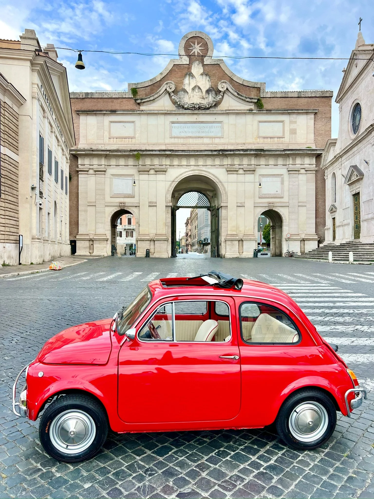 Piccolo carro rosso in stile vintage parcheggiato su strada di ciottoli davanti a un arco storico in una città europea.