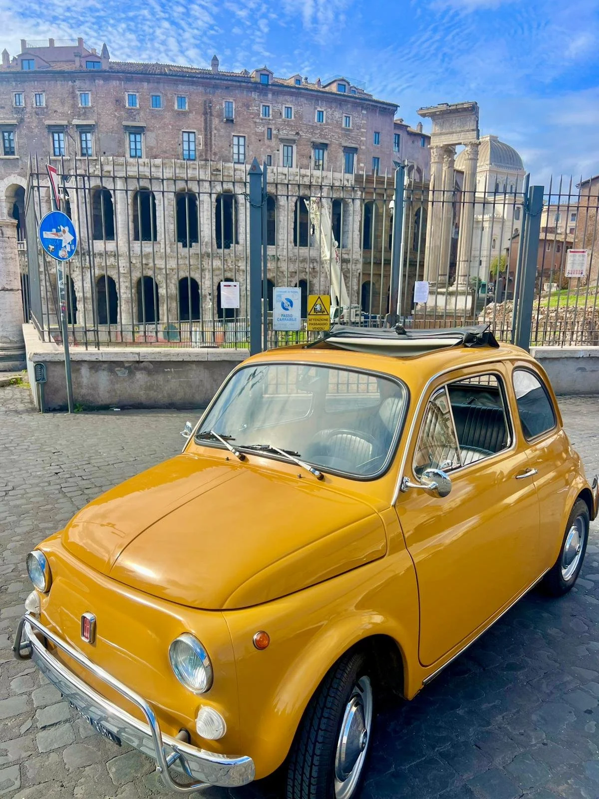 Auto d'epoca gialla parcheggiata su una strada di ciottoli davanti a un cancello in ferro e a un edificio antico.