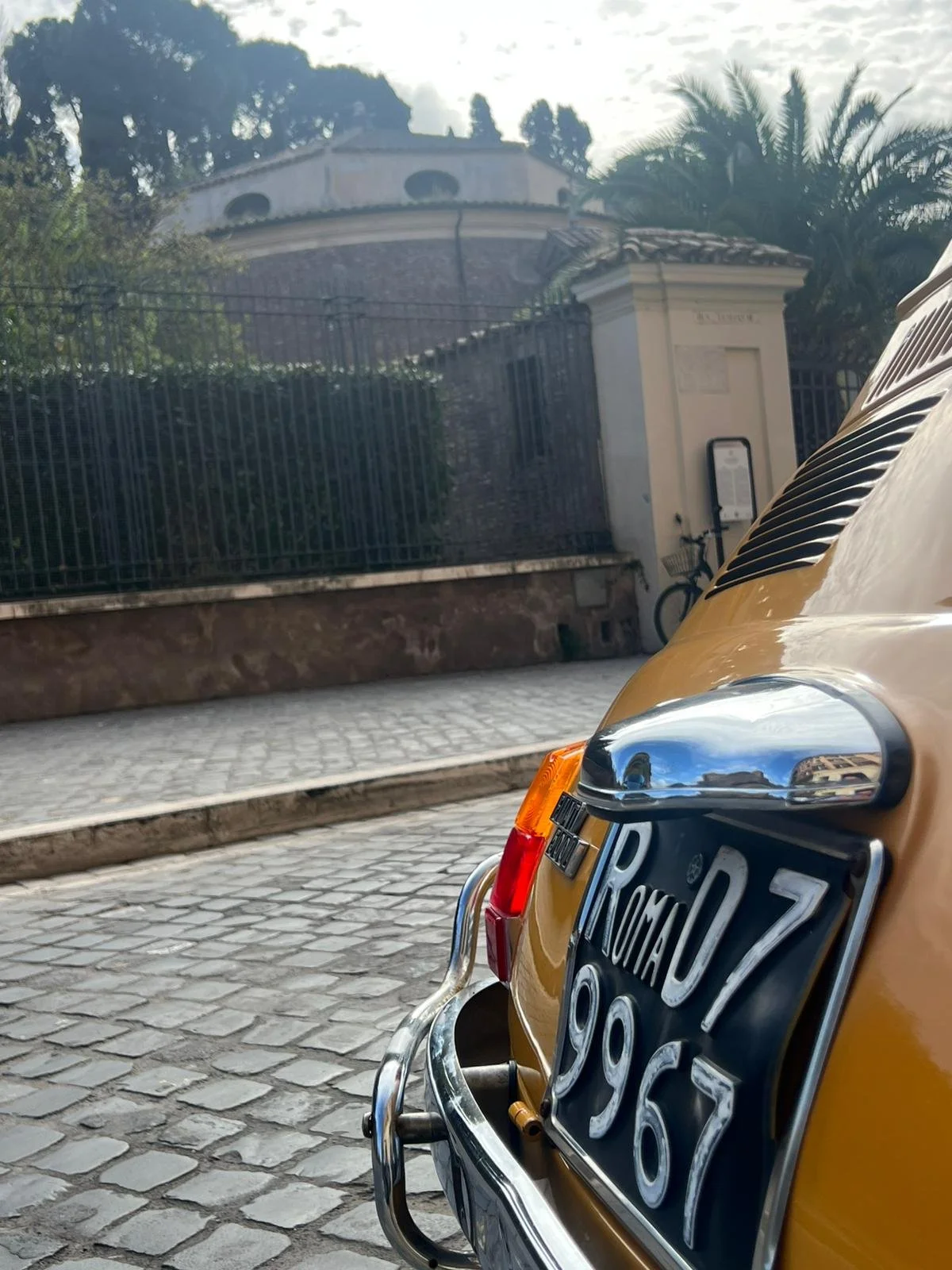Particolare di un'auto d'epoca di colore giallo con targa italiana, parcheggiata su una strada in ciottolato, con un edificio e alberi sullo sfondo.