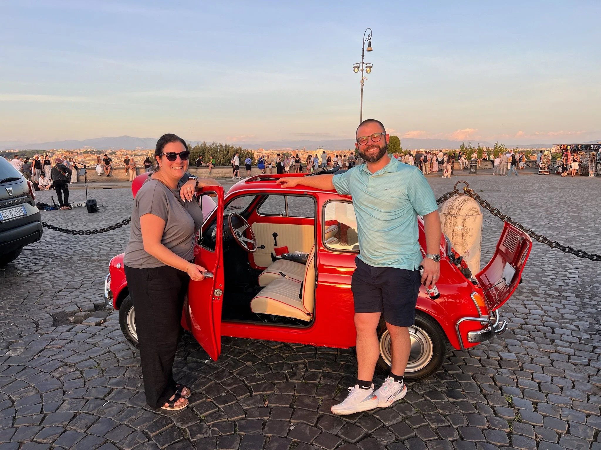 Una coppia felice con un'auto rossa vintage sulla passeggiata di sera, con molte persone in background e un tramonto sul fiume.