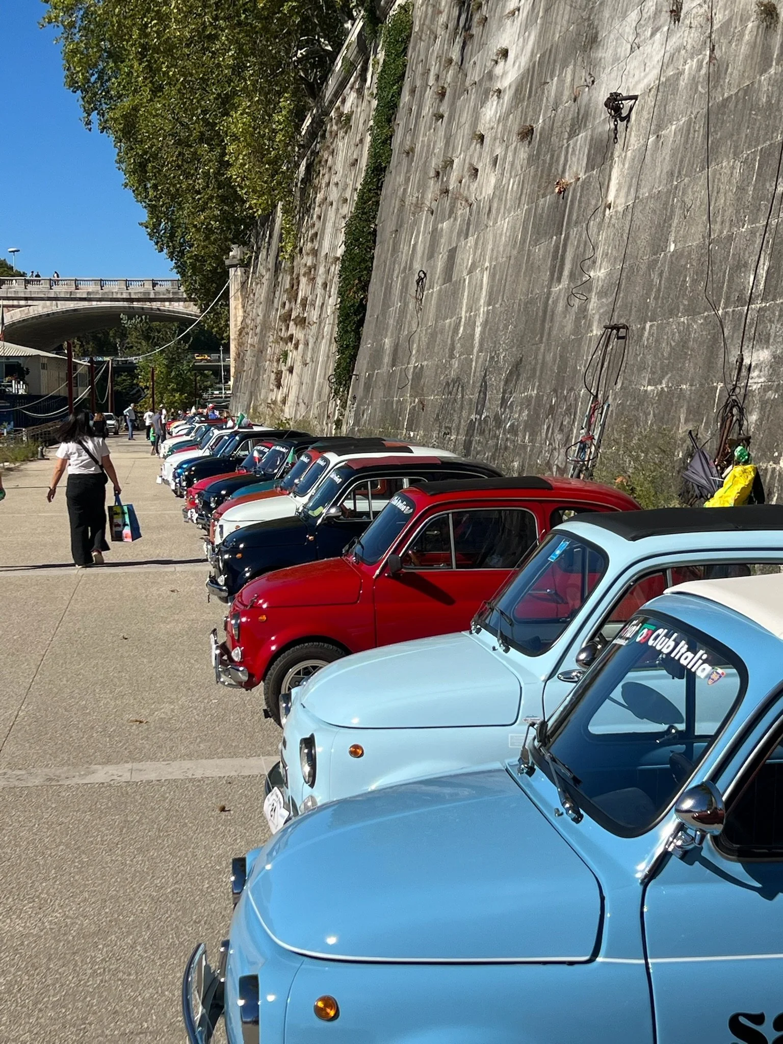File di piccole automobili classiche di diversi colori parcheggiate lungo un muro di pietra, con alcune persone che camminano nelle vicinanze.