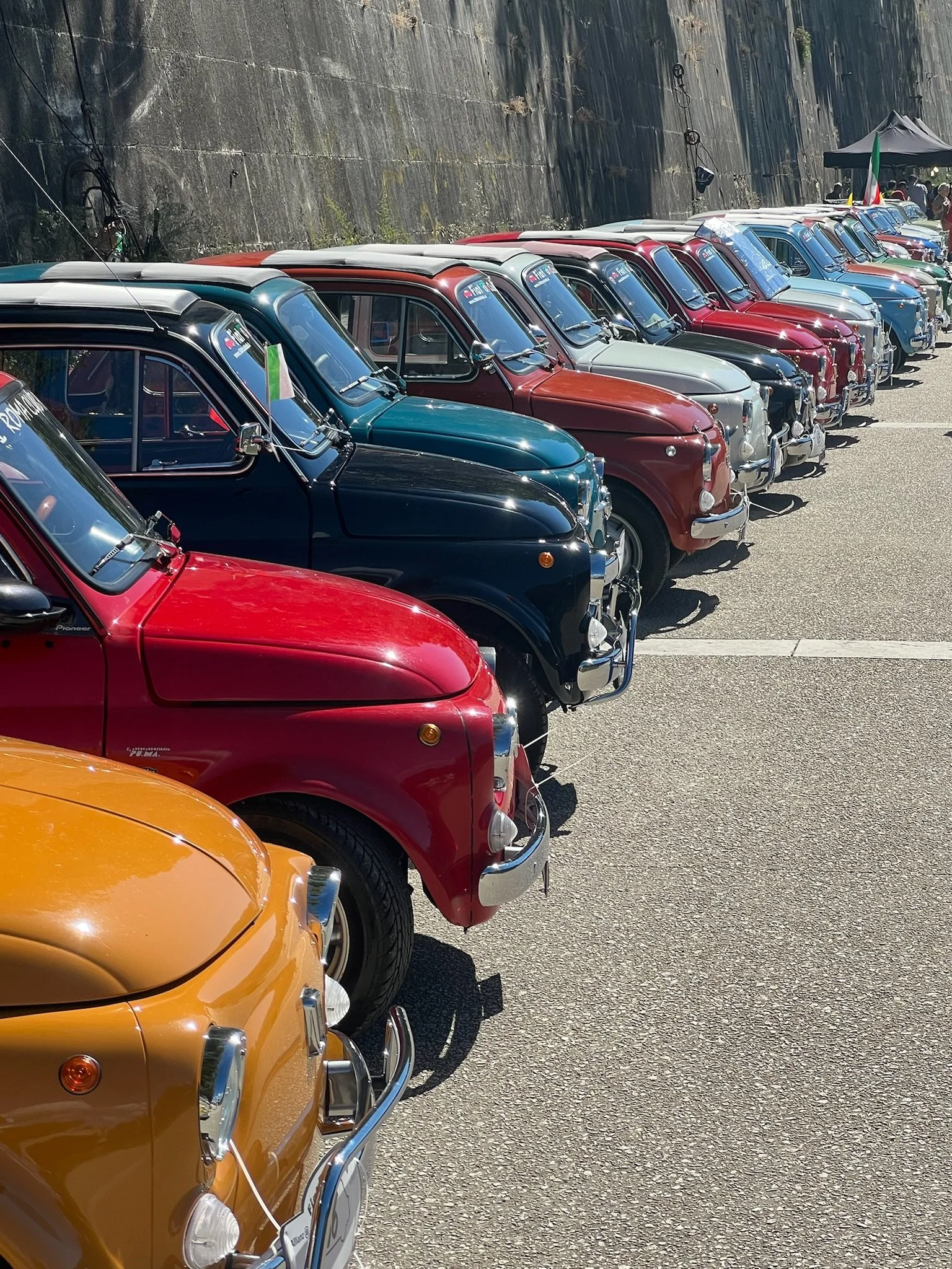 Riga di automobili d'epoca colorate parcheggiate lungo un muro di pietra, con alcune persone e tende sullo sfondo.