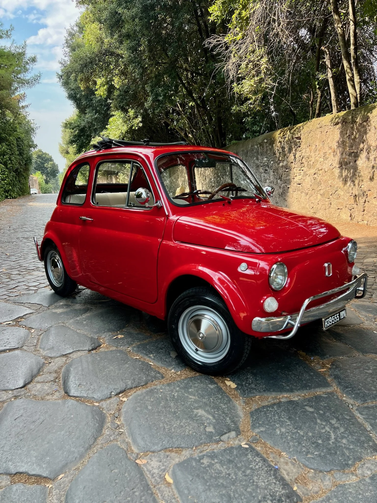 Auto d'epoca rosso parcheggiata su una strada di pietra con alberi e muro sullo sfondo.