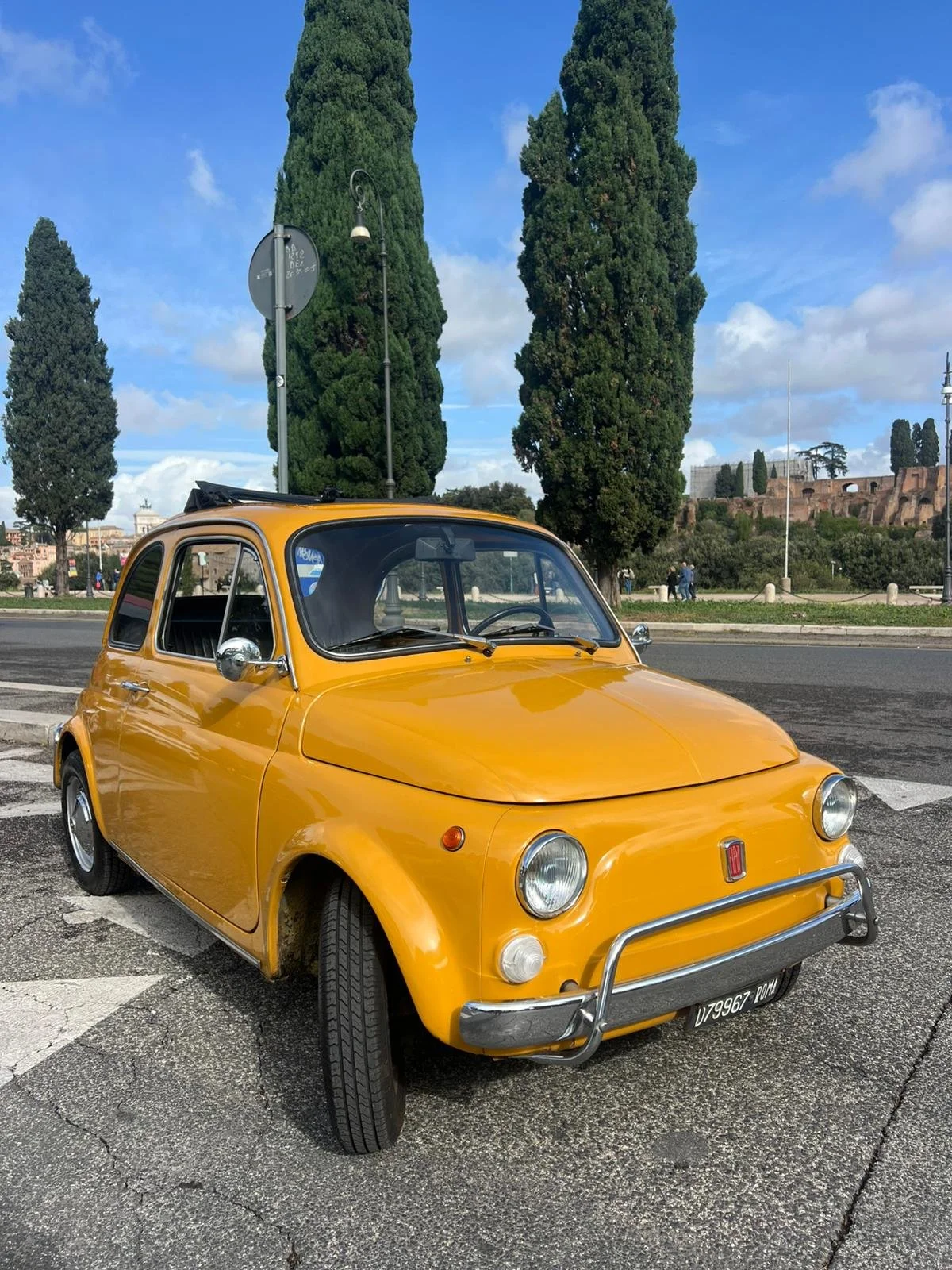 Auto d'epoca di colore giallo parcheggiata su strada con palazzi e alberi sullo sfondo.
