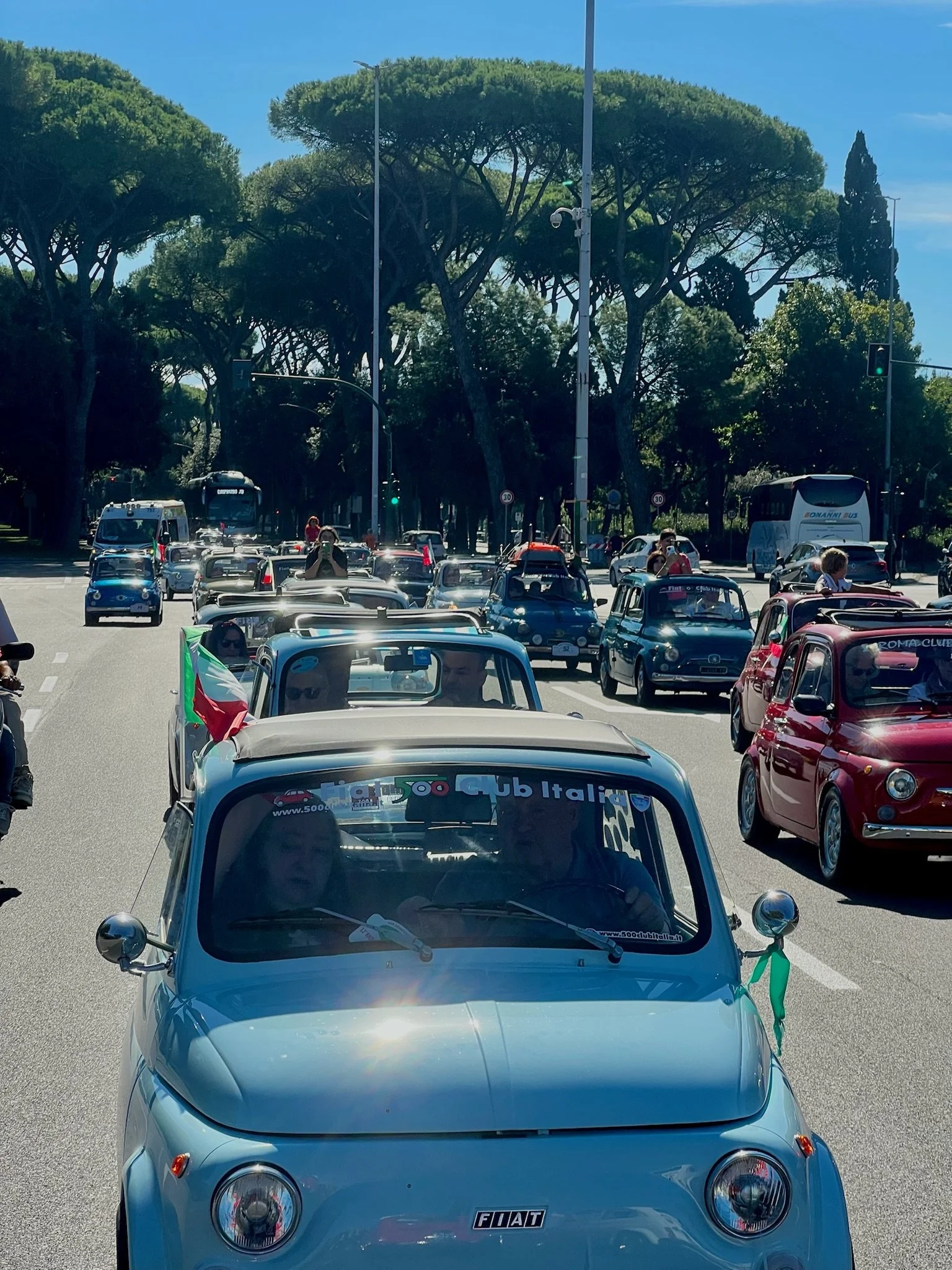 Una parata di auto d'epoca Fiat 500 che attraversano una strada alberata sotto un cielo sereno.