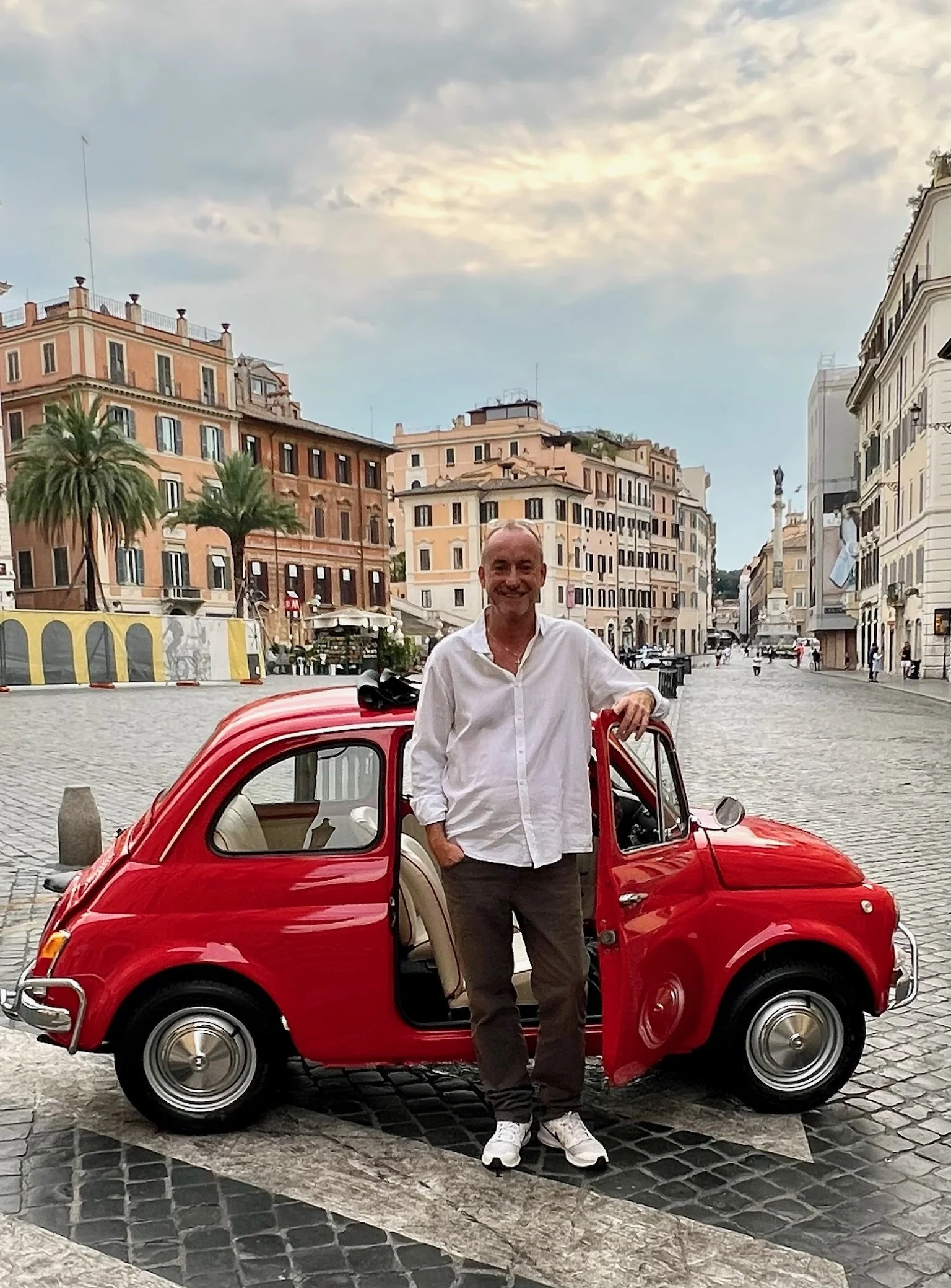 Un uomo sorridente vestito in camicia bianca e pantaloni scuri si trova accanto a una piccola auto rossa d'epoca in una piazza di città con edifici antichi e palme sullo sfondo.