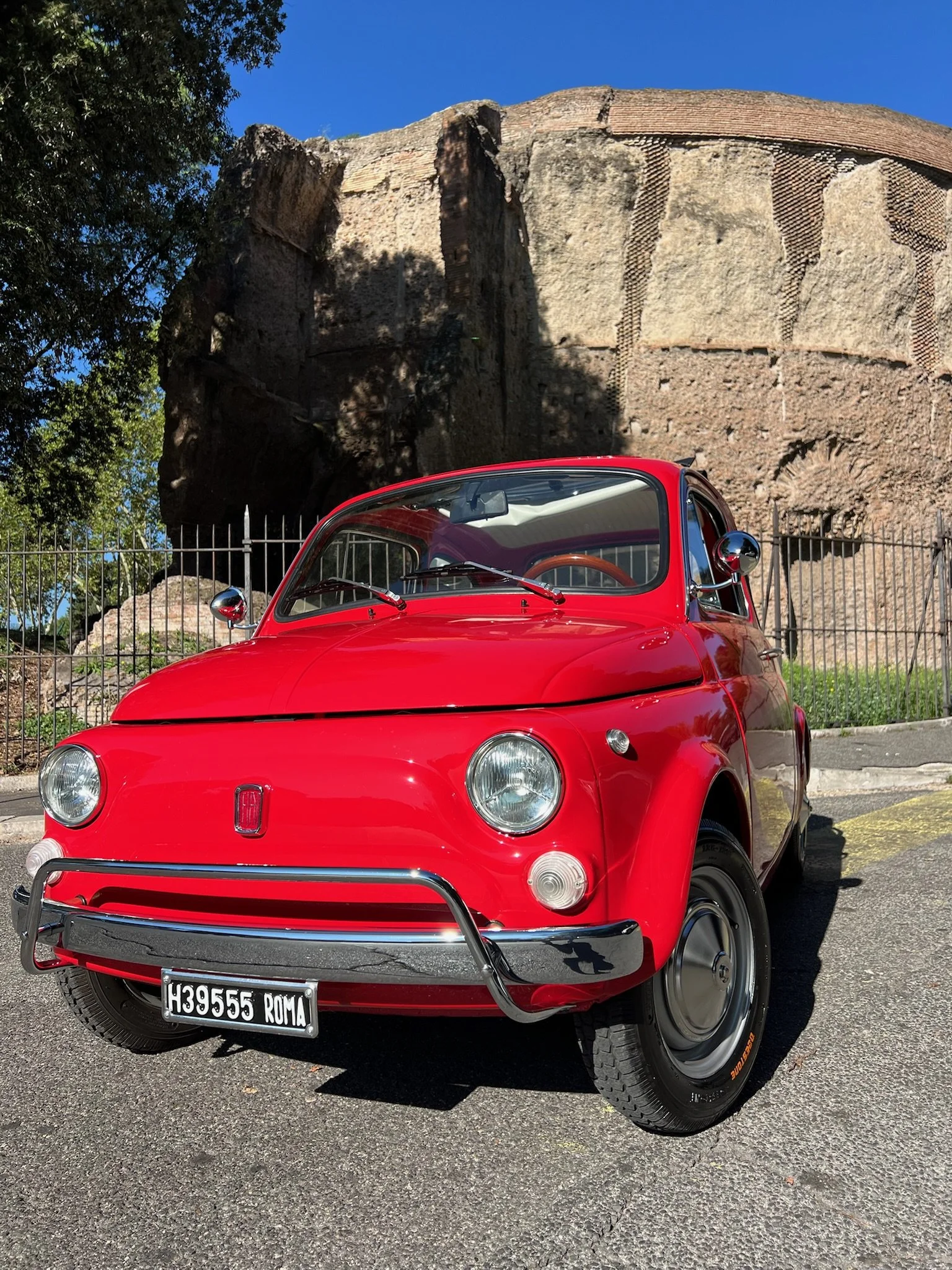 Auto rossa d'epoca parcheggiata su strada con sfondo di un'antica struttura in pietra e un cielo azzurro.