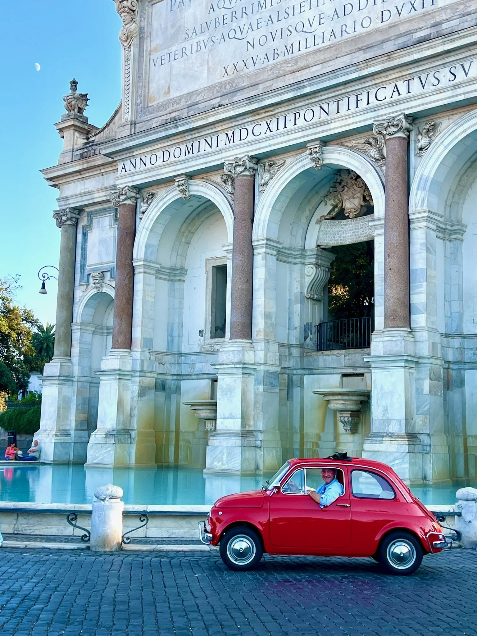 Un uomo seduto nel primo piano di una macchina rossa d'epoca, con un edificio storico in stile barocco sullo sfondo, con dettagli architettonici elaborati e iscrizioni in latino, e una fontana con acqua e alcune persone che siedono sulla panchina vic