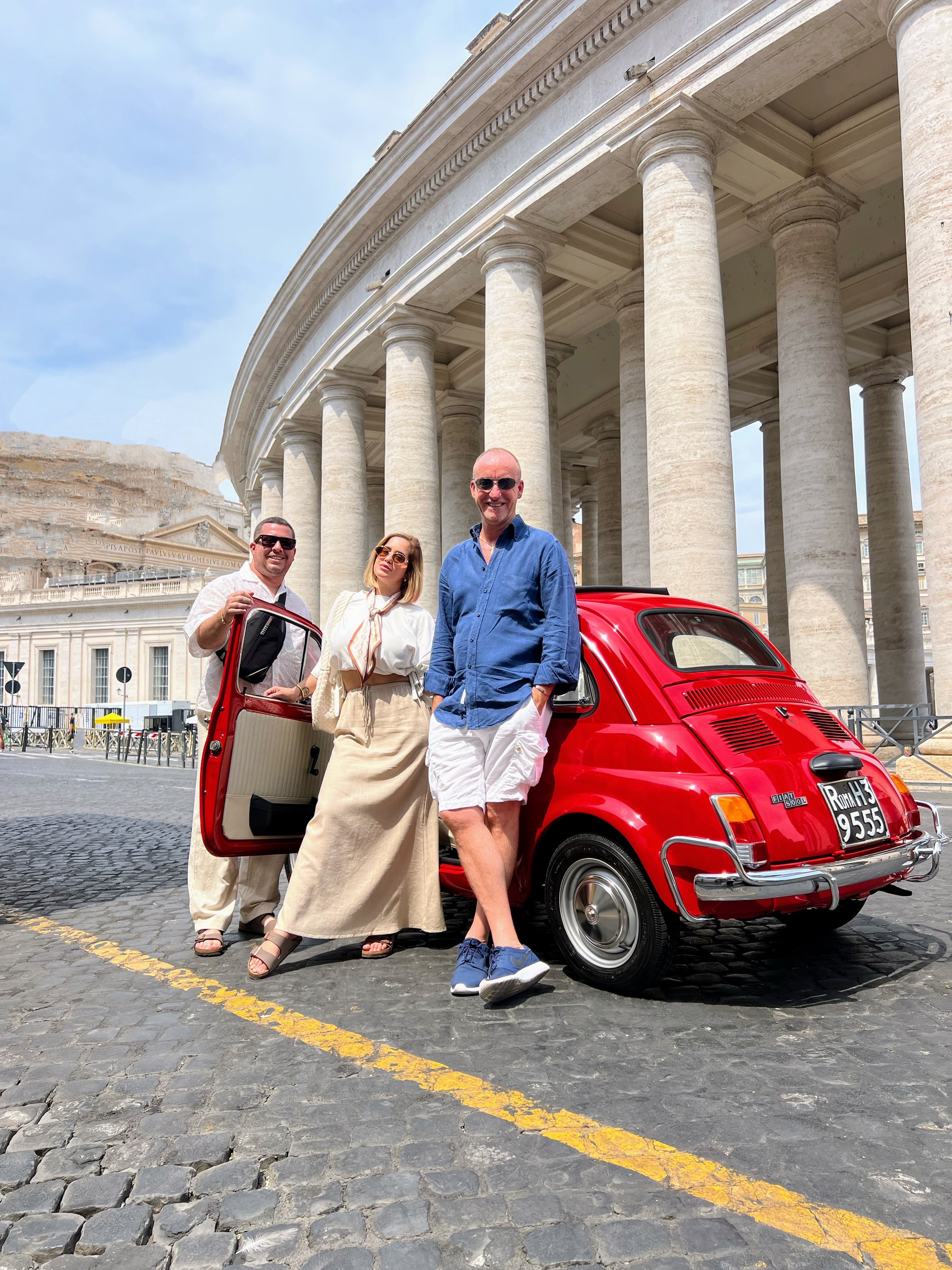 Tre persone posano davanti a una macchina rossa, con un edificio storico con colonne in background.