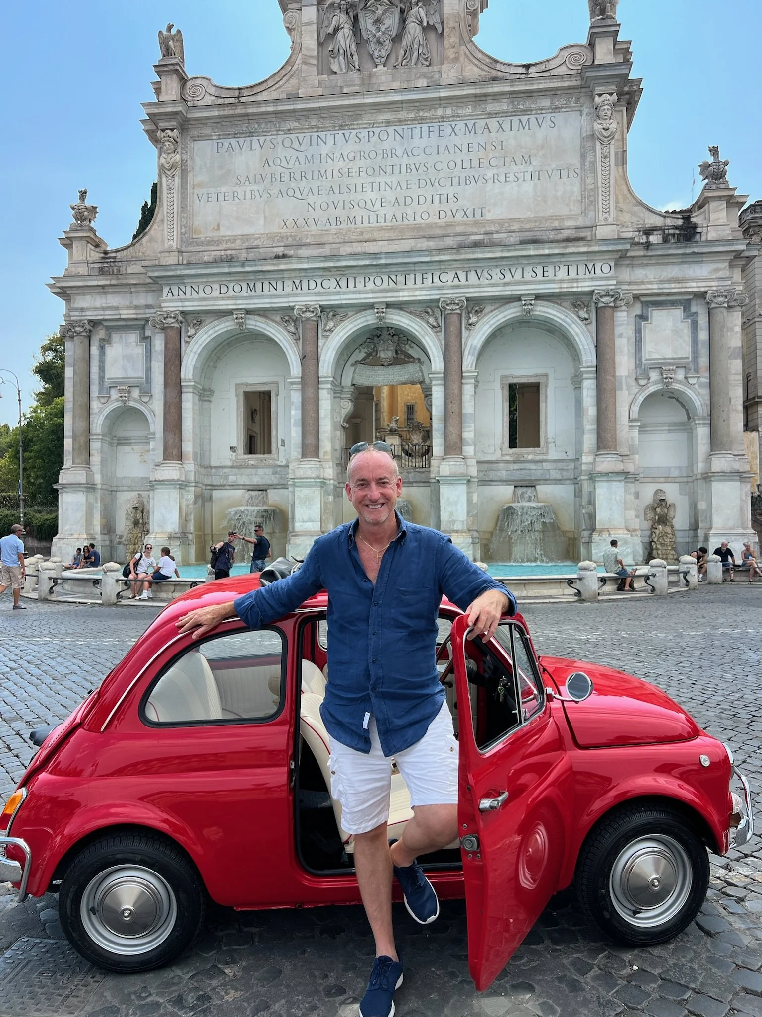 Uomo sorridente posa con una mini auto rossa davanti a una fontana e un edificio storico con iscrizione in latino.