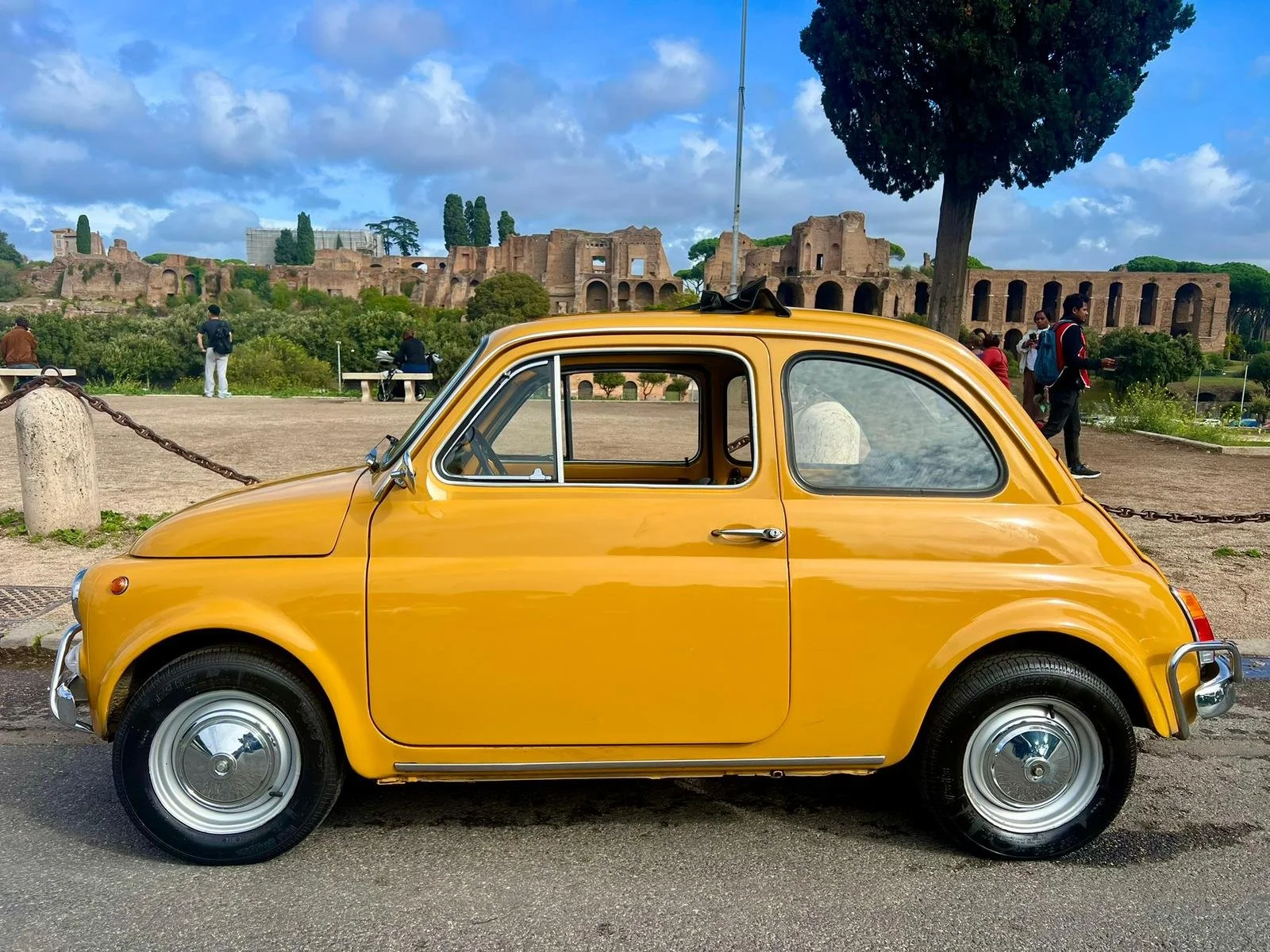 Una macchina d'epoca gialla parcheggiata su una strada con le rovine di un antico sito archeologico sullo sfondo e alcune persone intorno.