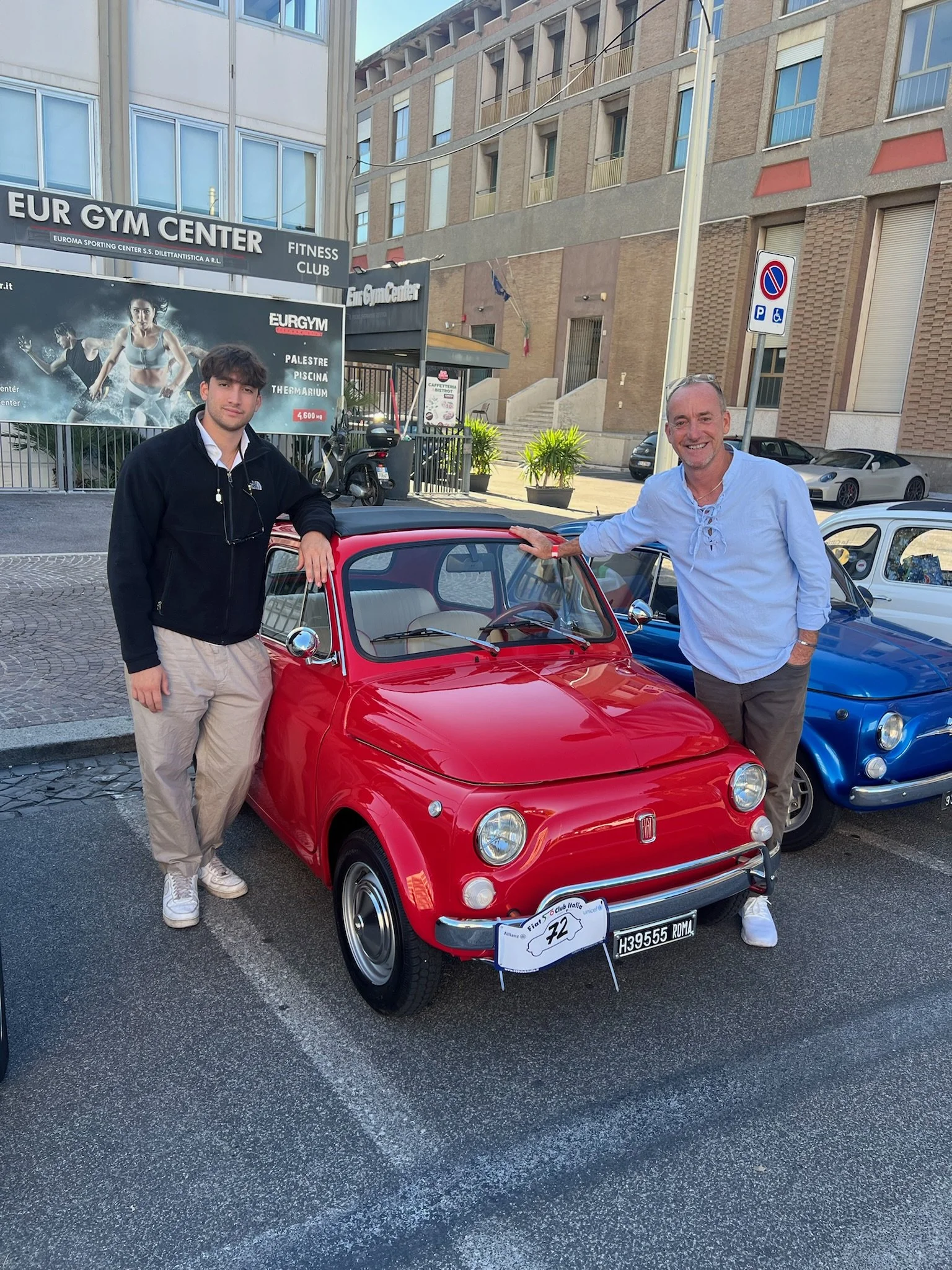 Due uomini davanti a una macchina d'epoca rossa, uno con camicia bianca e l'altro con felpa nera, in una piazza di città.