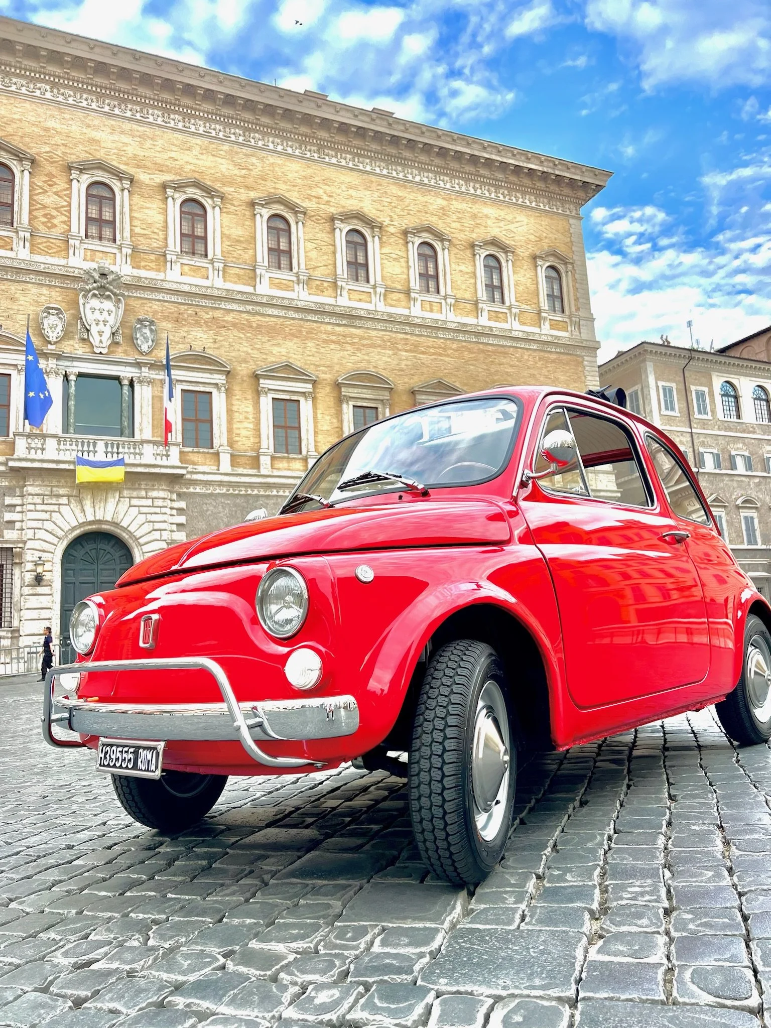 Auto rossa d'epoca parcheggiata su una strada in pietra, con un edificio storico sullo sfondo e un cielo azzurro con nuvole.
