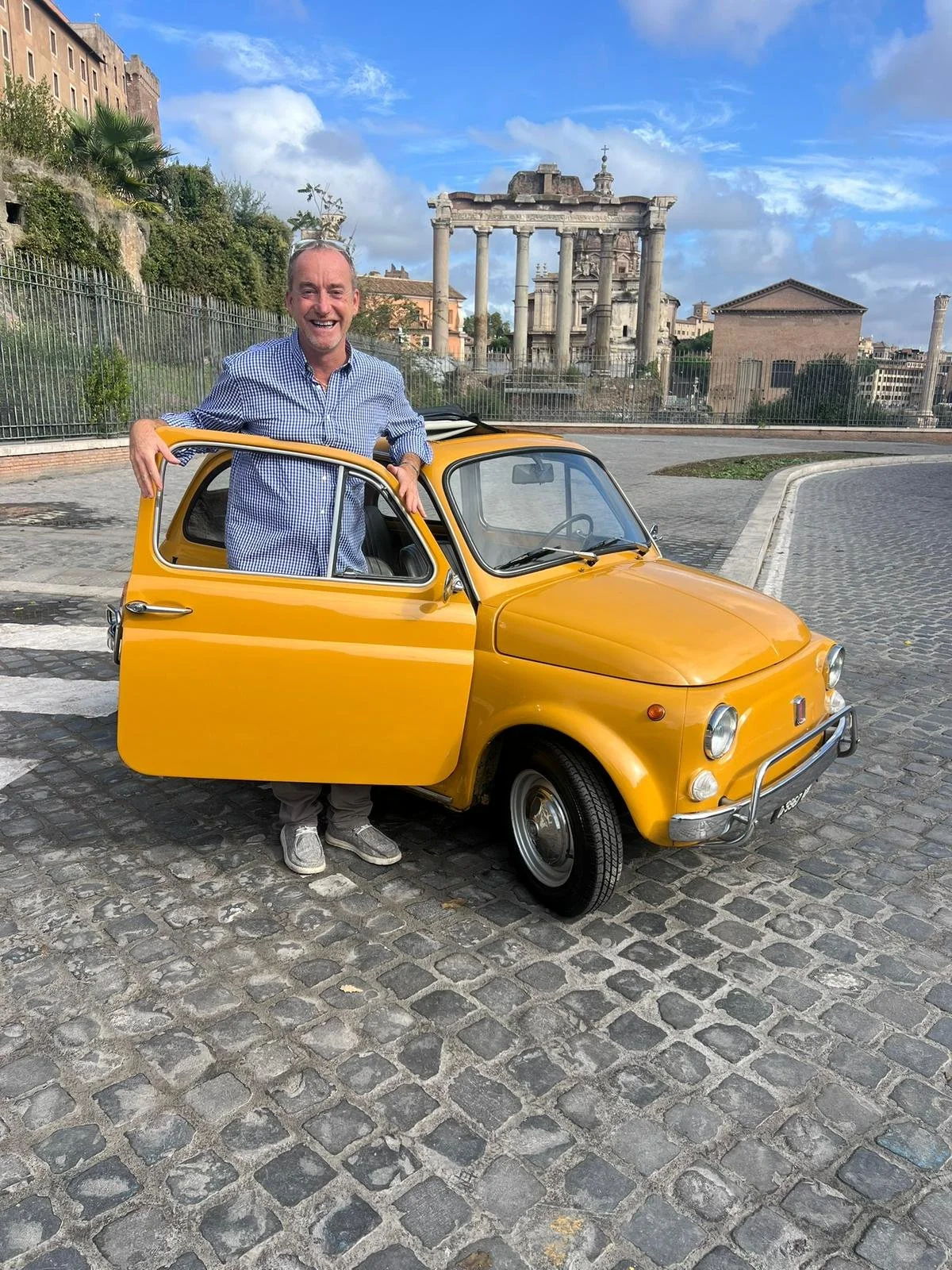 Uomo felice con camicia checkered blu, che si appoggia su una piccola auto gialla d'epoca, parcheggiata su una strada con ciottoli, sullo sfondo resti archeologici romani e un cielo parzialmente nuvoloso.