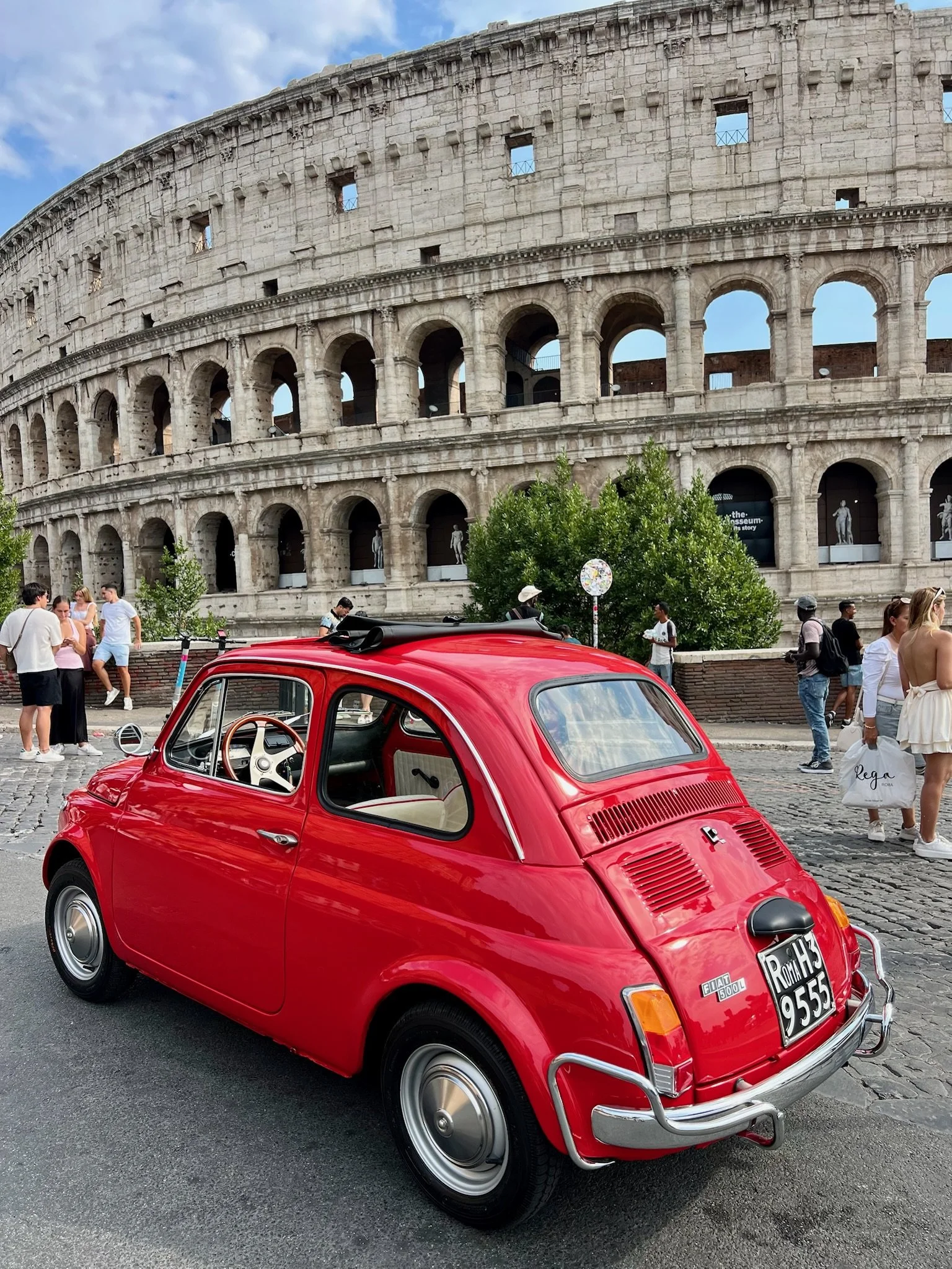 Una piccola auto rossa davanti al Colosseo a Roma, con alcune persone che passeggiano e si divertono.