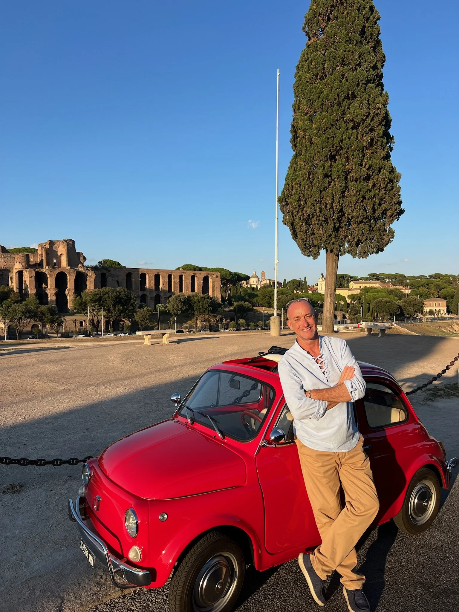 Uomo sorridente che si appoggia a una piccola auto rossa in un'area all'aperto con architetture antiche sullo sfondo, al tramonto.